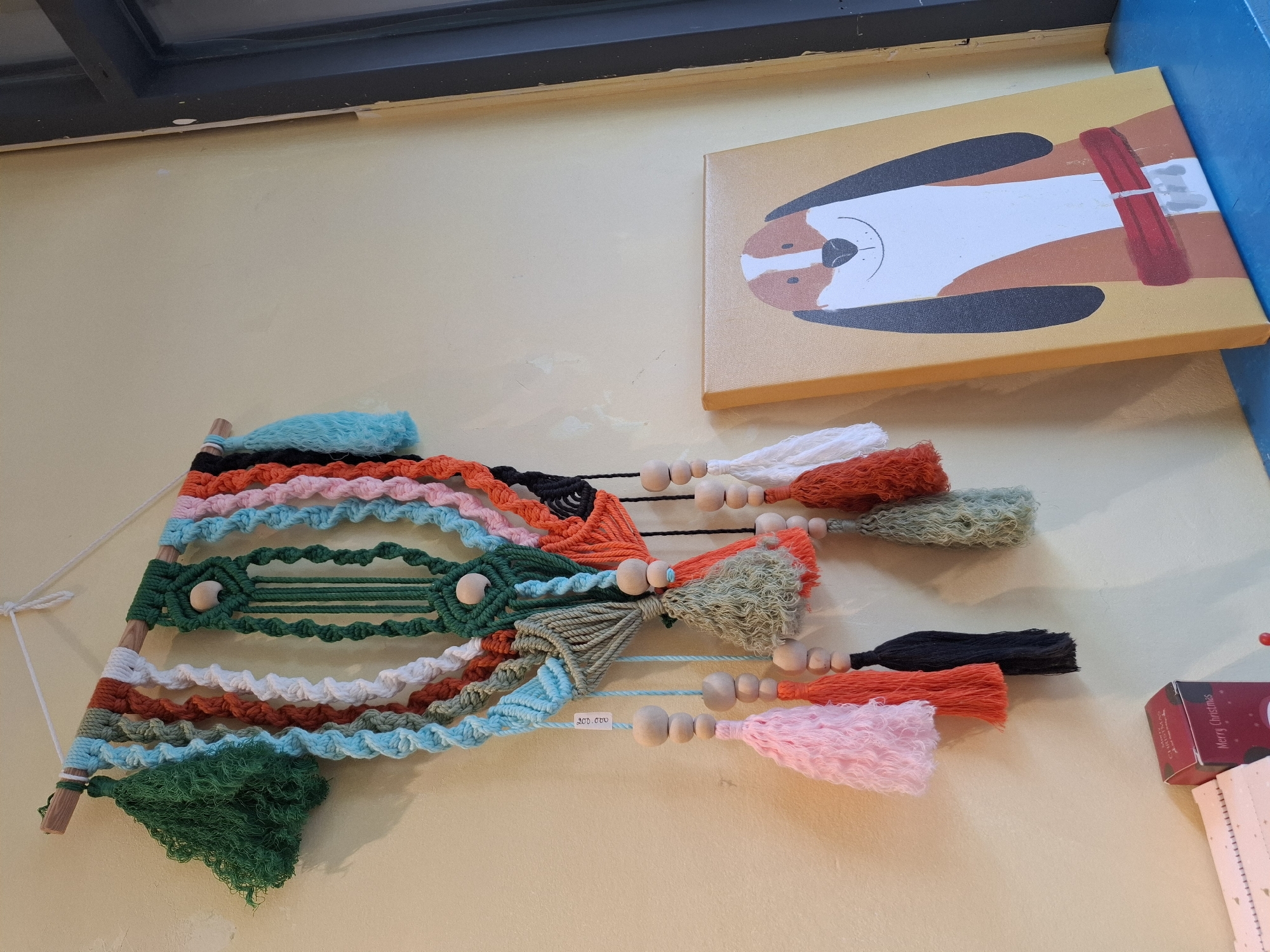 The image shows a macrame wall hanging with multiple colorful yarn strands and wooden beads, suspended from a wooden dowel. Below the wall hanging, on a blue surface, is a small canvas painting of a dog with brown and white markings and a red collar, with a yellow background. To the left of the painting, a red box with "Merry Christmas" written on it is visible. The background is a plain yellow wall, with a dark vertical element, likely a door or window frame, on the right side of the image. The setting appears to be an indoor space, possibly a shop or a home. The location is not specified within the image.