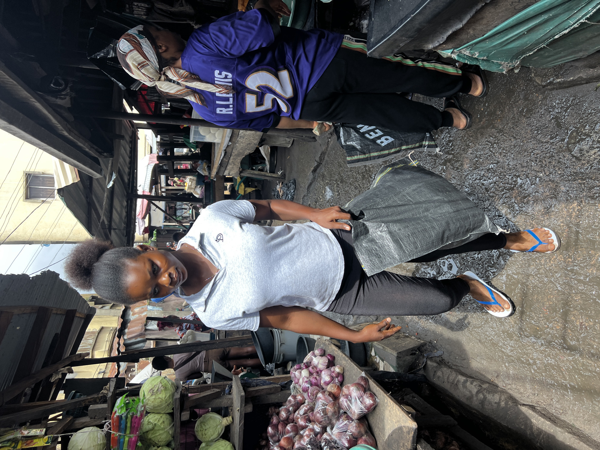 This image depicts a bustling street market in Nkpor, Nigeria. A young woman, with dark skin and her hair styled in a high bun, walks towards the viewer, carrying a large black bag. She is wearing a light gray polo shirt and dark leggings, accessorized with white flip-flops with blue straps. She is looking downwards with a focused expression. To her right, another individual wearing a purple jersey with "R. LEWIS 52" on the back and a patterned headscarf is visible from the back, holding a light-colored object.
The market environment is characterized by makeshift stalls constructed with corrugated iron and wood. In the foreground, to the left, is a wooden stall displaying large heads of cabbage and bags of red onions. The ground is wet and muddy, suggesting recent rain or cleaning. In the background, other market stalls and people can be seen, hinting at the vibrant commercial activity of the location. Power lines are visible overhead, typical of urban Nigerian settings.