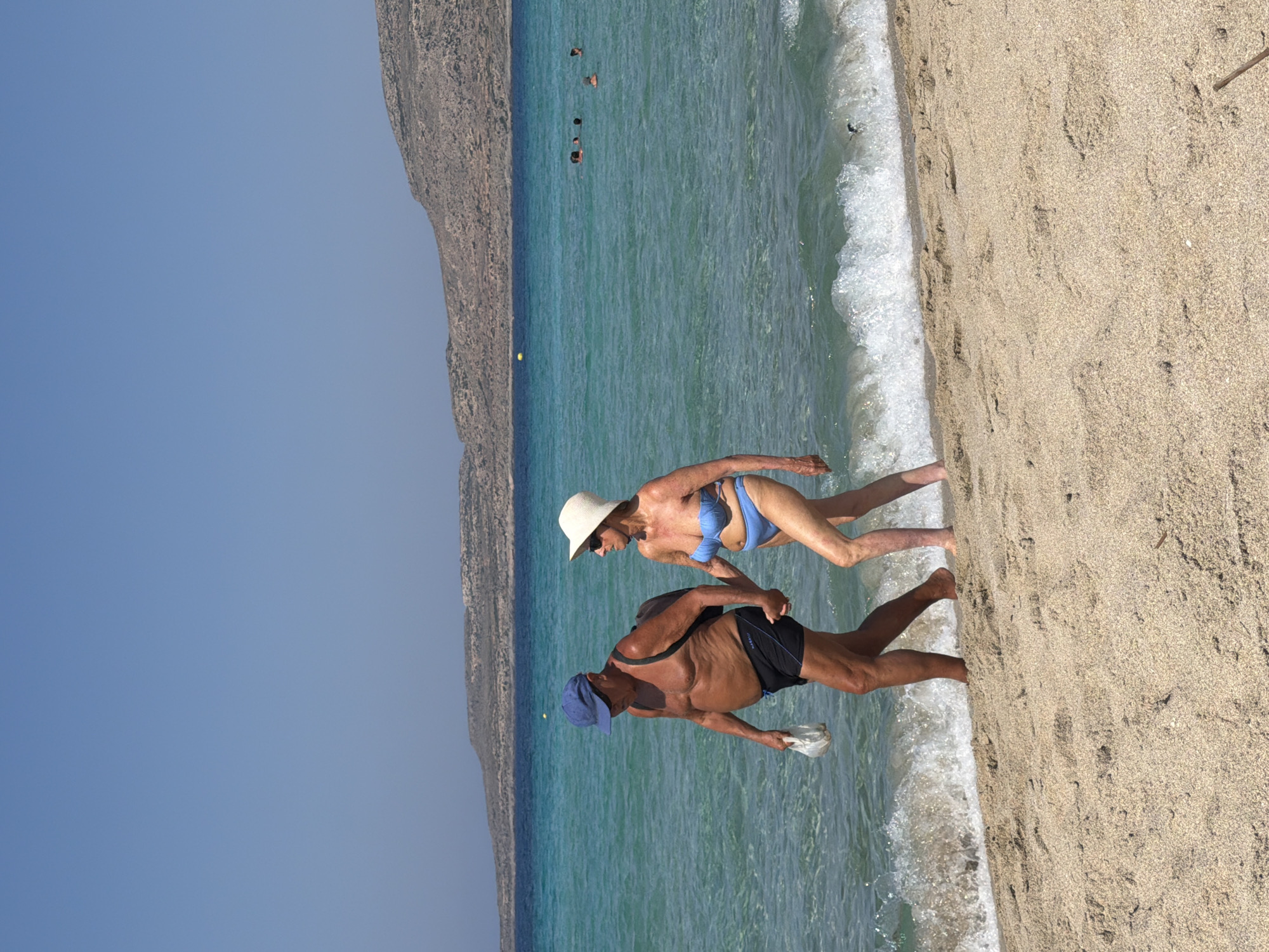 An elderly couple walks hand-in-hand along a sandy beach in Greece. The man, wearing a blue baseball cap and black swim trunks, carries a white plastic bag in his left hand. The woman, wearing a white hat and a blue bikini, walks beside him. Gentle waves lap the shore, and other beachgoers are visible in the turquoise water in the background. Mountains rise in the distance under a clear blue sky.