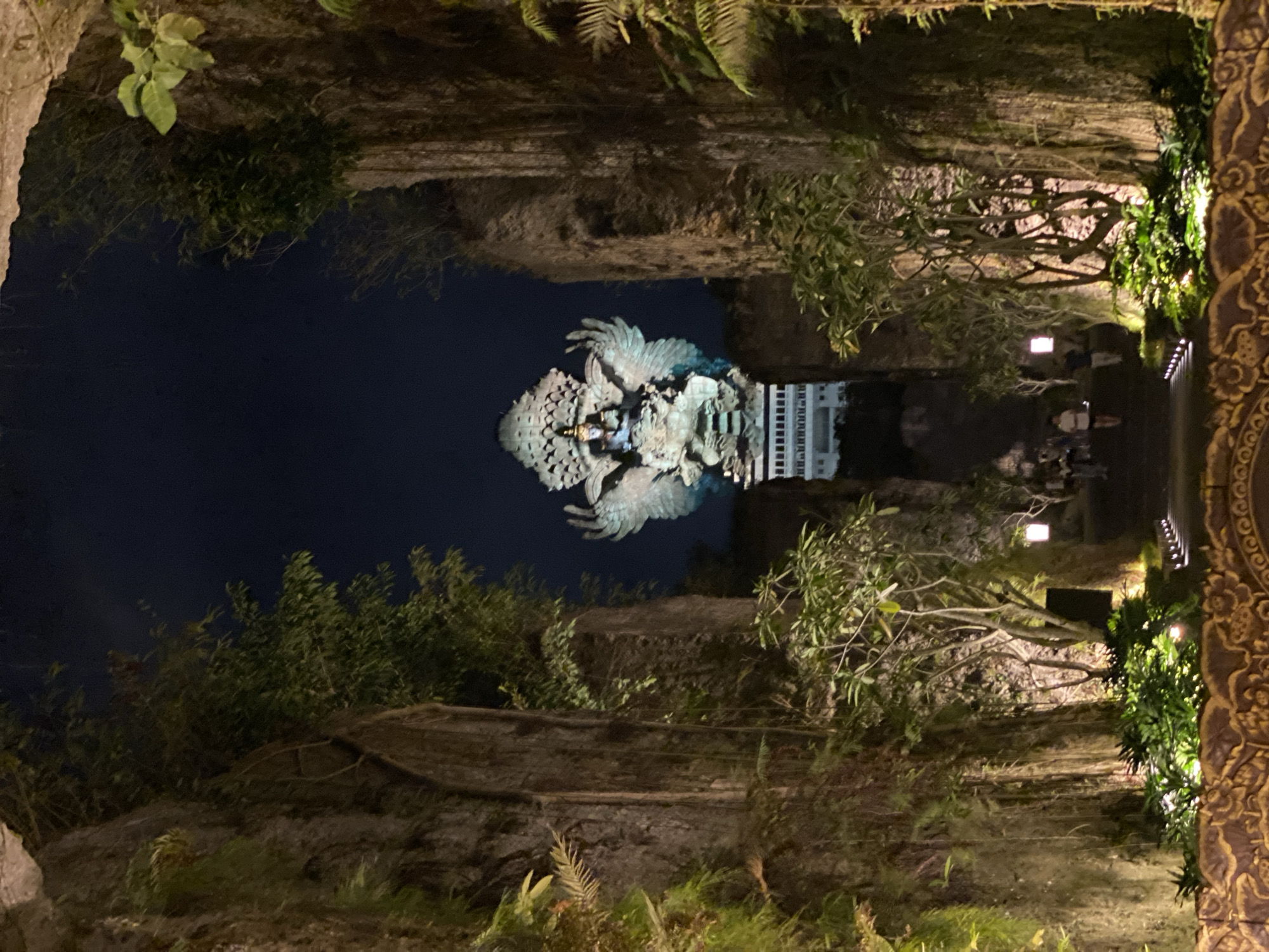 The image depicts the Garuda Wisnu Kencana (GWK) Cultural Park in Uluwatu, Kuta Selatan, Indonesia, at night. A colossal statue of Garuda Wisnu, a Hindu deity, dominates the background, illuminated against the dark sky. The statue, a prominent landmark, features a bird-like creature with outstretched wings, seated on a pedestal.
The foreground is framed by towering, rough-textured rock walls on either side, overgrown with lush green vegetation and vines. Pathways illuminated by soft lighting wind through the scene, leading towards the statue. Several individuals are visible walking along these pathways, appearing to be visitors or patrons of the park. The environment is characterized by natural rock formations, dense foliage, and artificial lighting, creating a dramatic and atmospheric setting. The overall scene suggests an outdoor cultural or tourist attraction experienced after sunset.