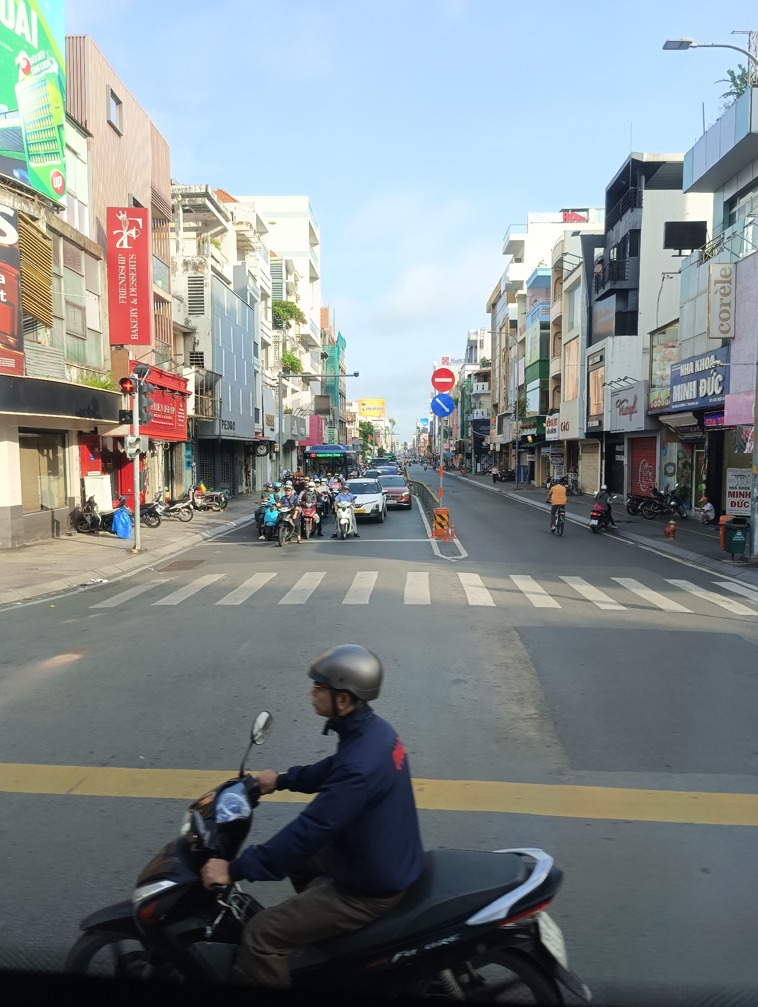 This is a daytime, eye-level, vertical, medium shot of a street scene in Thủ Đức City, Vietnam. The street is lined with multi-story buildings featuring various businesses. On the left side of the street, a red building with "FRIENDSHIP BAKERY & DESSERTS" signage is visible. Traffic is present, including numerous motorcycles, a bus, and a white car. A pedestrian crossing is marked on the asphalt road. In the foreground, a person wearing a dark blue jacket and a silver helmet is riding a dark-colored motorcycle. The sky is clear with a light blue hue.