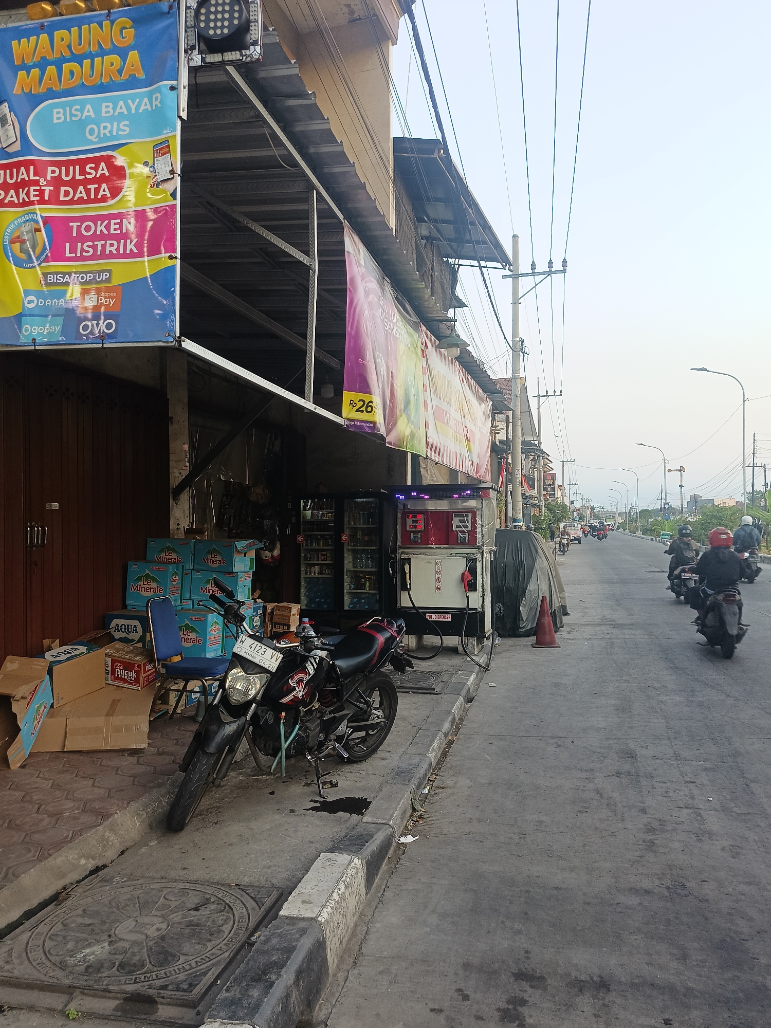 The image shows a street scene in Gedangan, Indonesia. On the left side of the road, there is a shop with a banner that says "WARUNG MADURA" and advertises payment options like QRIS. The shop has a brown wooden roll-up door. In front of the shop, there are stacks of bottled water, a blue chair, and a motorcycle parked on the sidewalk. A fuel dispenser is also visible next to the shop. The street is paved and has a curb. Several motorcycles are seen driving on the road, with riders wearing helmets. There are power lines overhead. The sky is bright, indicating daylight.