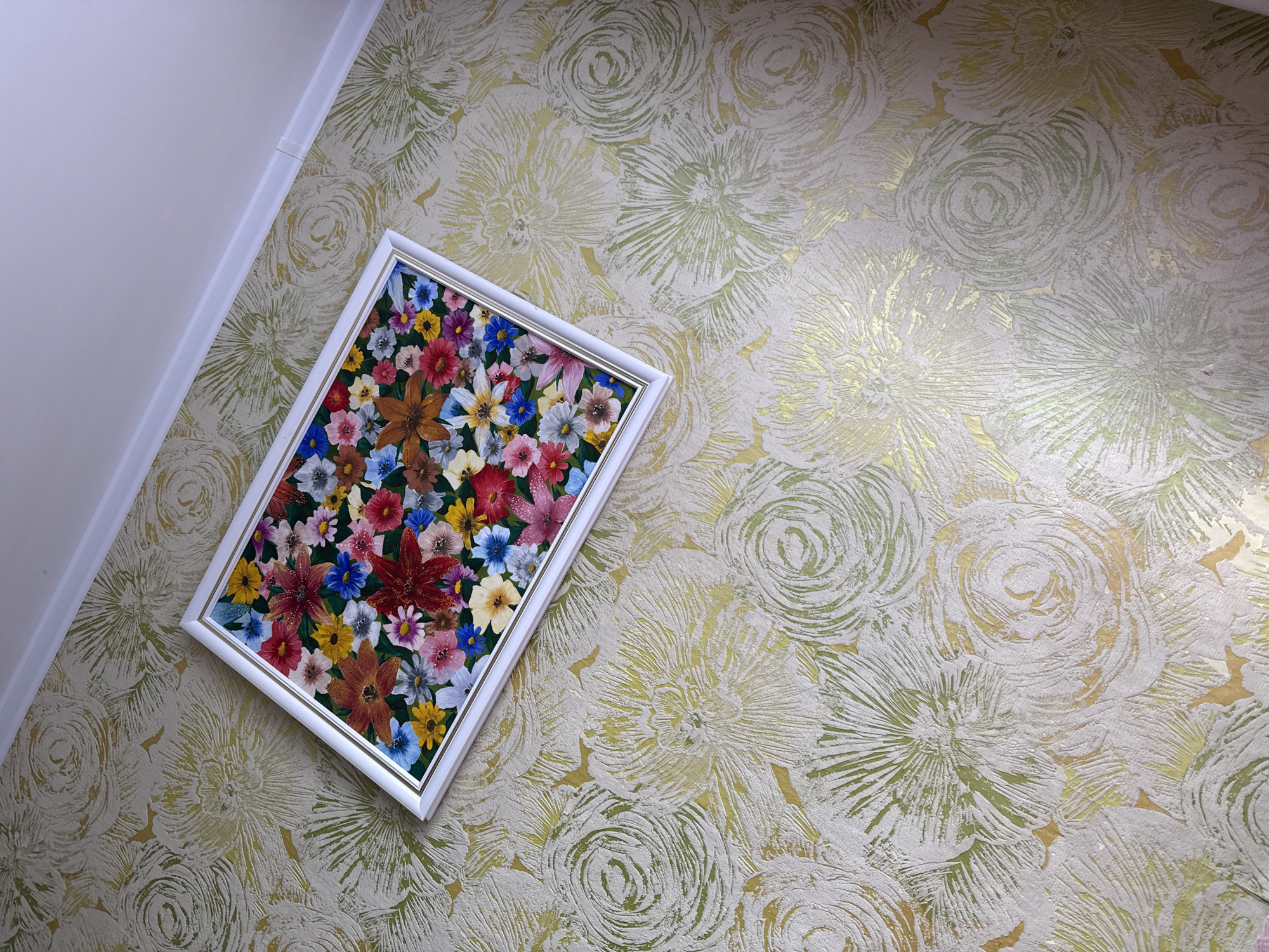 An interior shot captures a framed painting of a floral arrangement resting on a textured wallpapered surface. The painting features a vibrant medley of flowers in various colors including red, pink, blue, yellow, and white. The wallpaper has a repeating pattern of large, stylized floral motifs in muted tones of green, gold, and off-white, with a distinct textured finish. The scene is lit from above, casting soft shadows. The image is oriented to show the painting at an angle. The setting is an indoor residential space, possibly a living room or hallway, indicated by the white baseboard and wall junction visible in the upper right corner. The location is identified as Krasnogorskoye, Russia.