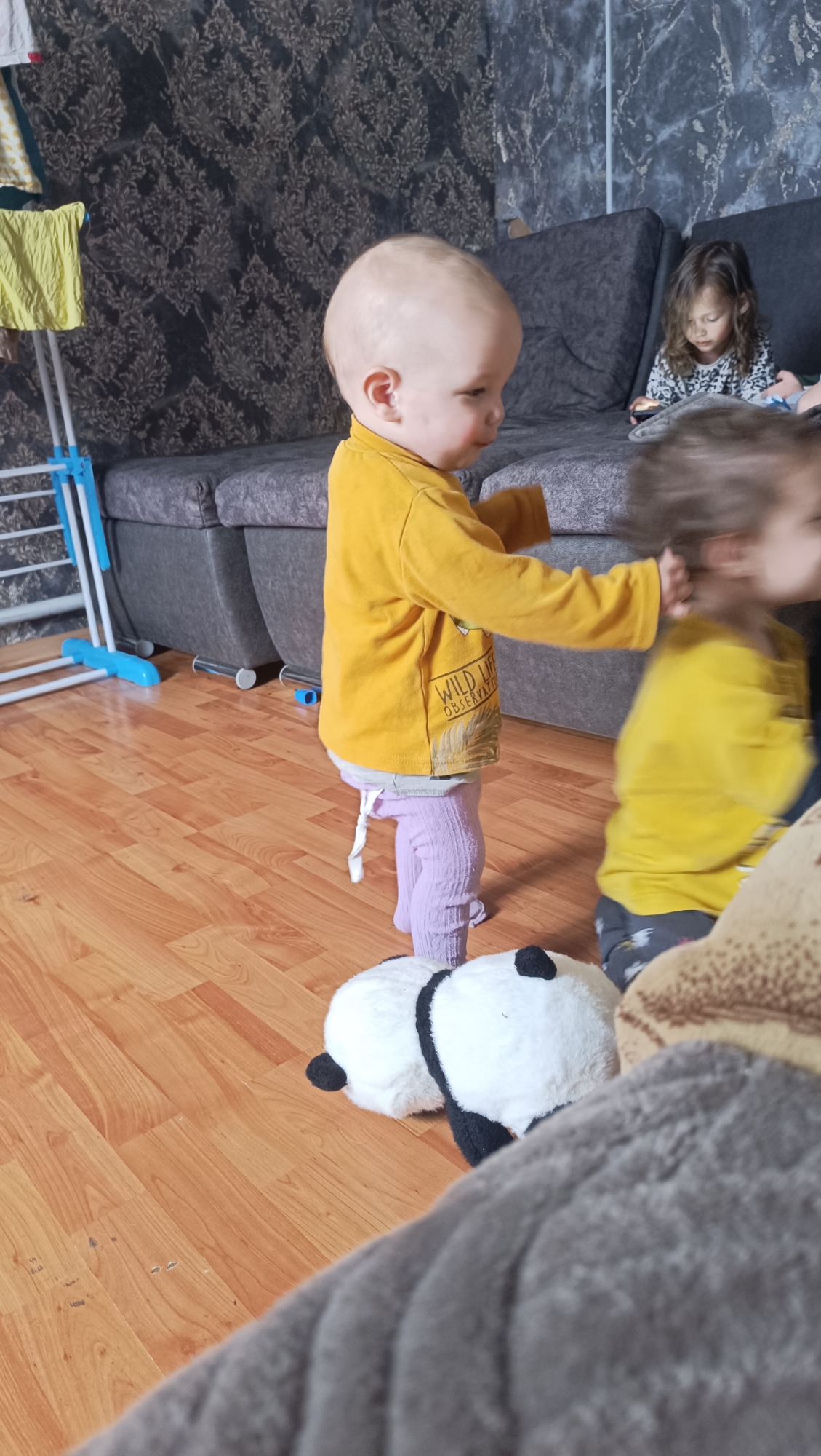 An infant, wearing a yellow long-sleeved shirt and purple leggings, stands on a wooden floor in a living room. The infant's head is turned to the right. The infant's hands appear to be reaching towards another child who is blurred and to the right of the frame, also wearing a yellow long-sleeved shirt. Two panda stuffed animals are on the floor in front of the infant. In the background, a couch is visible, with a young girl sitting on it and looking at a smartphone. The room has patterned wallpaper and a clothing rack with yellow clothing to the left. There is no indication of the city of Karsha, Russia.