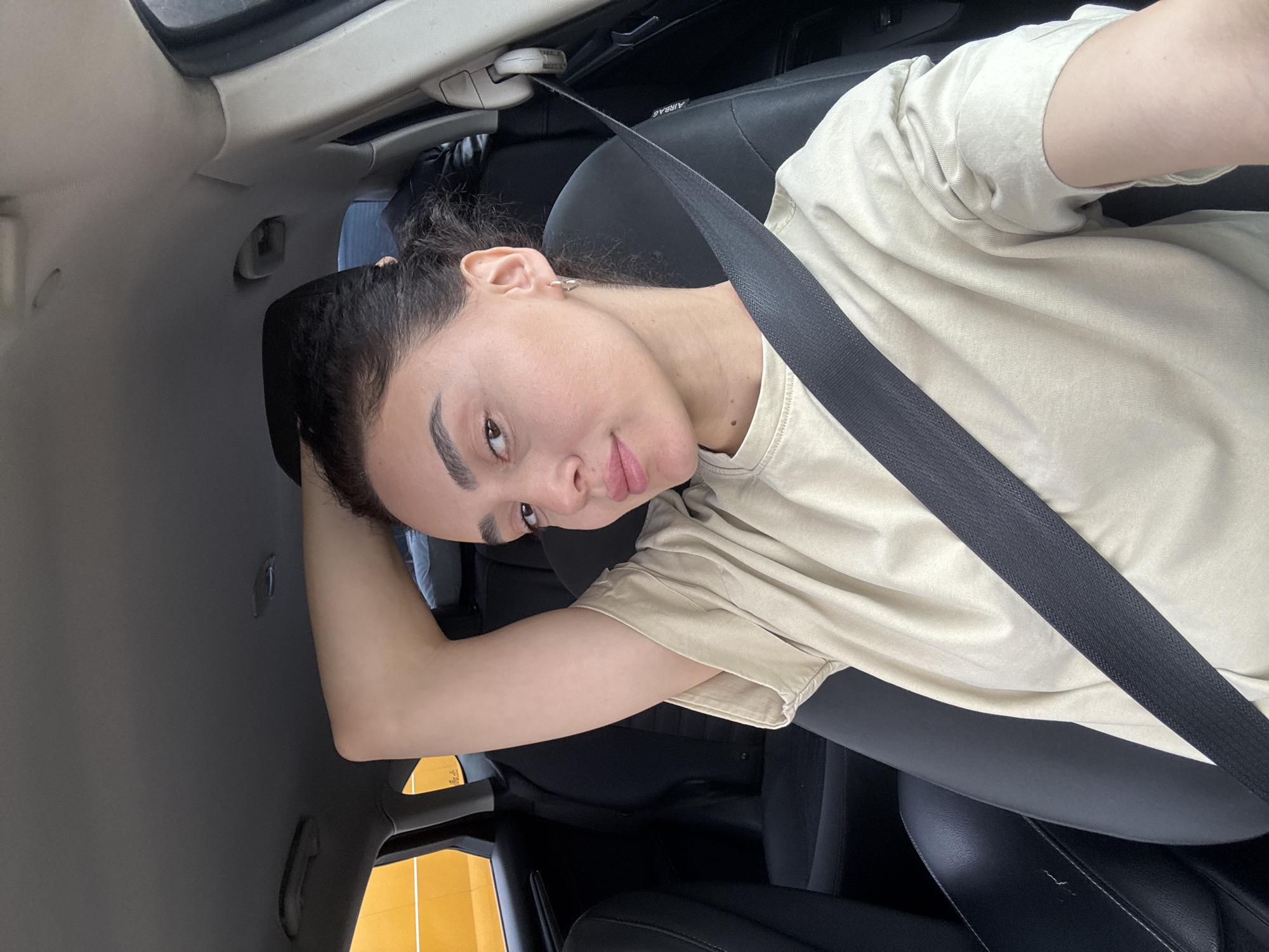 A woman is seated in the driver's seat of a car, wearing a seatbelt. She is facing the camera. She is wearing a beige t-shirt. The interior of the car appears to be black and gray. There are yellow panels seen outside the window. The location is Tbilisi, Georgia.