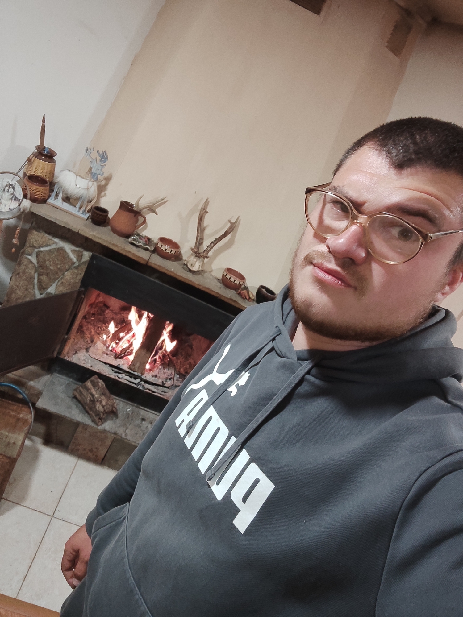 A man with brown hair, a beard, and glasses is taking a selfie in front of a stone fireplace with a fire burning inside. The fireplace mantel is decorated with various items, including pottery, antlers, and a small ceramic reindeer. The man is wearing a gray hooded sweatshirt with a white "PUMA" logo. The location is in a room with light colored walls. No specific evidence is provided that indicates that the picture was taken in the city of Varshilo, Bulgaria.