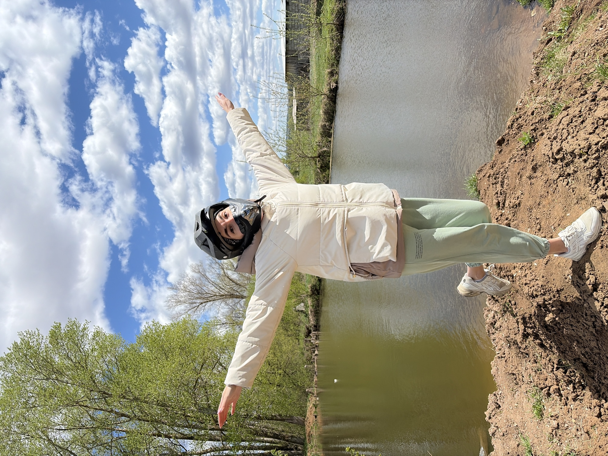 The image shows a person wearing a black helmet and a beige jacket standing on a dirt bank next to a body of water. The person is holding their arms out to the sides. The person is also wearing light green sweatpants and white sneakers. The environment includes trees, grass, and a blue sky with white clouds. The location is Nizhnii Vishur, Russia.