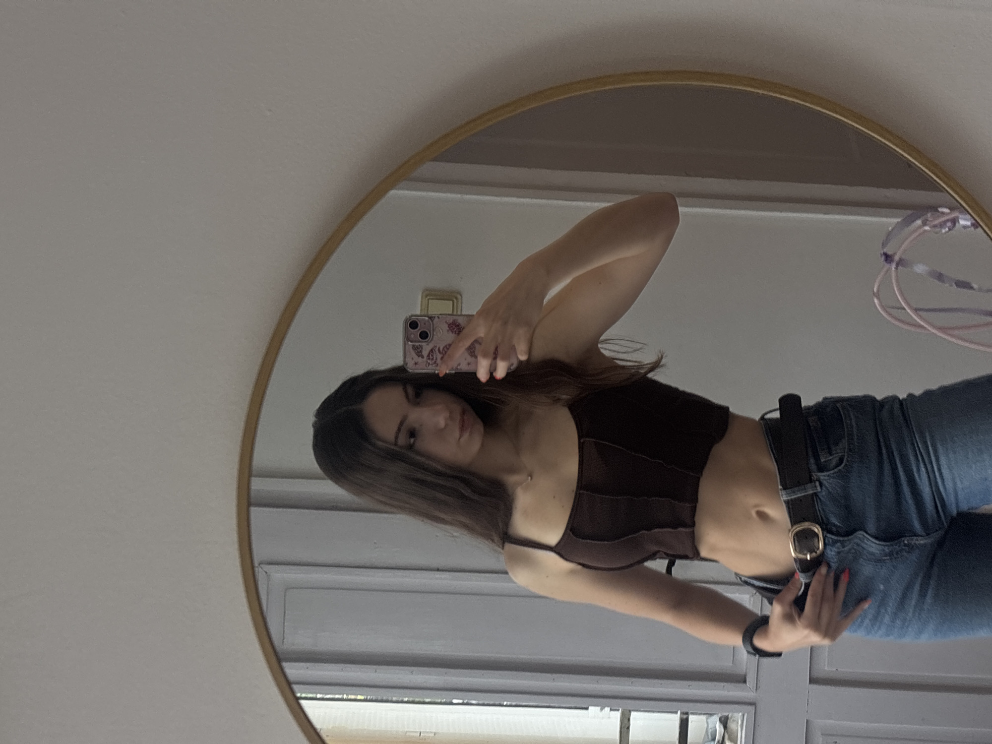 The image is a selfie taken in a mirror. A young woman with long brown hair is posing. She is wearing a dark brown top and blue jeans with a black belt. She is holding a phone in her left hand, taking the picture. A circular mirror with a gold frame reflects the scene. The background includes a window with white frames, and part of a light-colored wall. There are no indications or information to suggest it is taken in the city of Kaspichan, Bulgaria.
