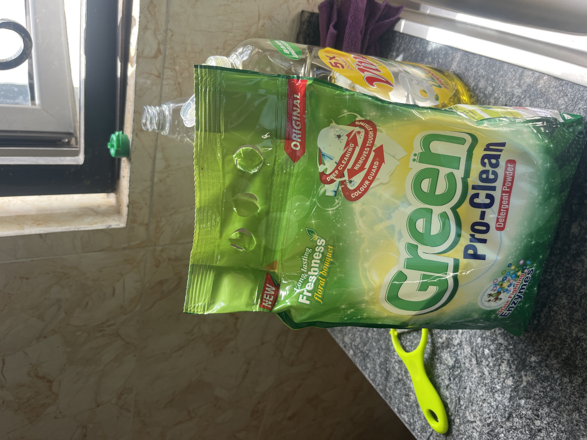 A green bag of Green Pro-Clean detergent powder is placed on a speckled gray countertop next to a clear plastic bottle of dish soap. A lime green peeler is to the left of the detergent bag. In the background, a window with a dark frame lets in natural light, illuminating the scene.