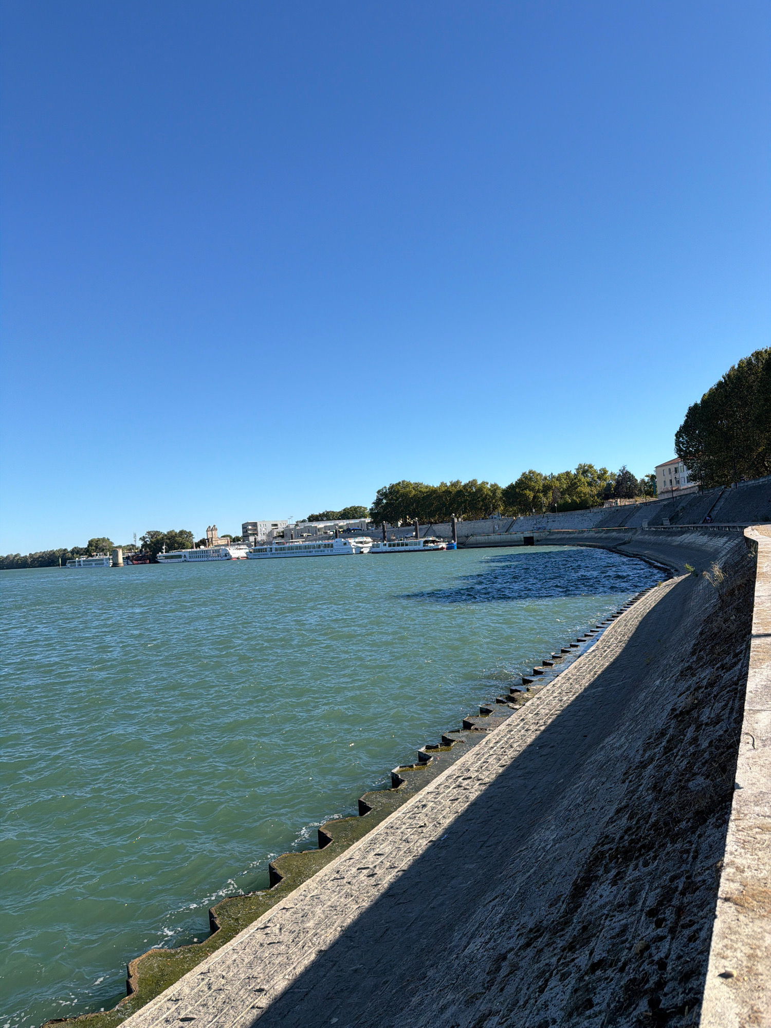 This is a photograph of the Rhone River embankment in Arles, France. The image captures a bright, sunny day with a clear blue sky. The water of the river is a greenish-blue hue and appears slightly choppy. Along the riverbank, there is a curved, stone embankment. Steps are integrated into the embankment, descending into the water in places. Several river cruise ships are docked on the far side of the river, with buildings and trees lining the opposite bank. The embankment in the foreground is casting a distinct shadow across the water and the lower part of the embankment itself. There are no people visible in the image.