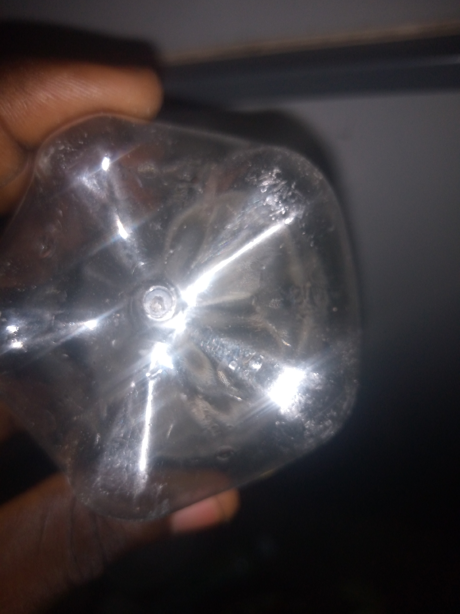 The image shows the bottom of a clear plastic bottle, likely a water bottle, held by a person's hand. The bottom of the bottle has a star-shaped pattern of indentations. There are some small bubbles or imperfections visible on the plastic. The background is dark and out of focus, with a hint of a horizontal line suggesting a shelf or surface. The scene appears to be indoors, with artificial lighting reflecting off the plastic. There is no indication of the location being in Jauro Yinu, Nigeria.