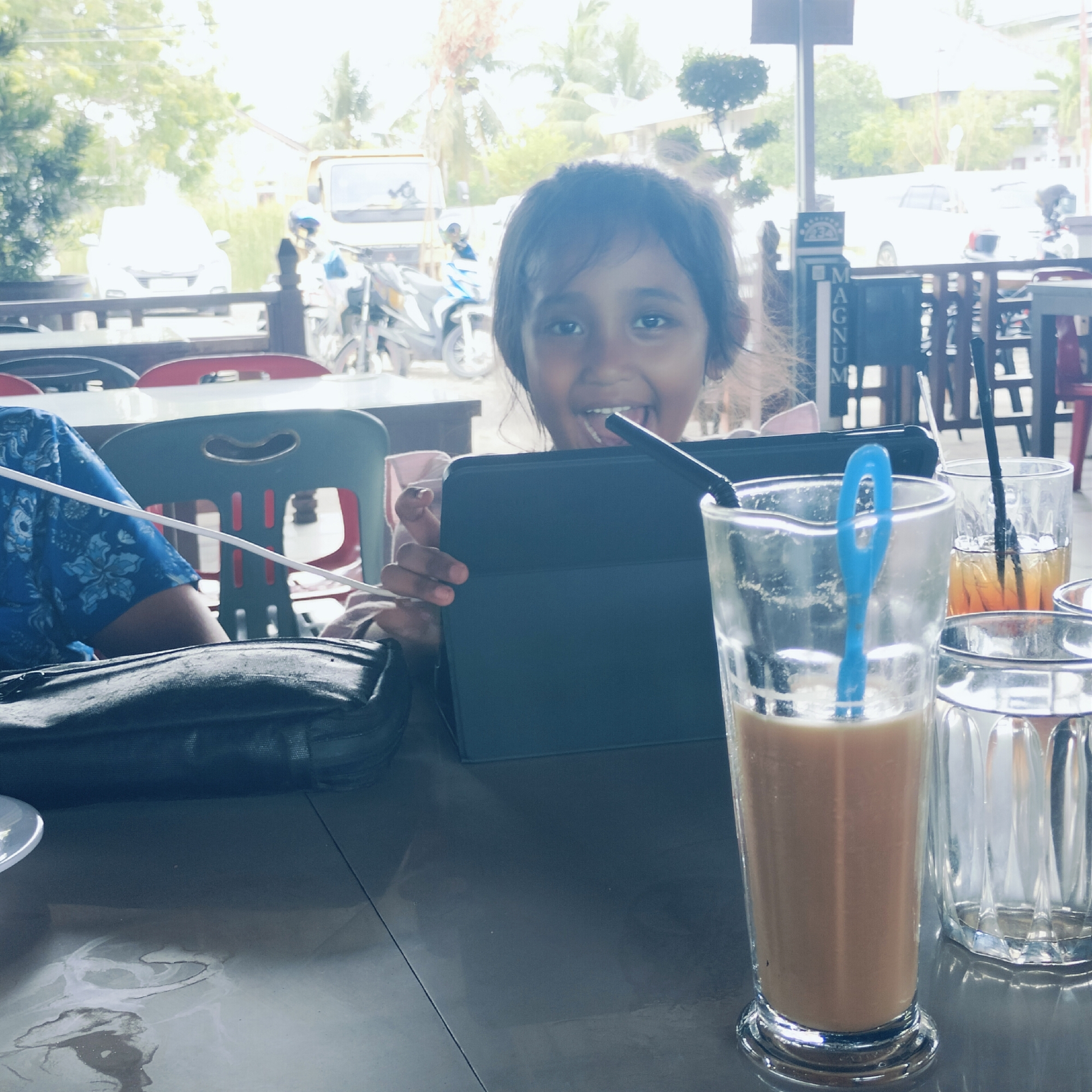 A young girl, with dark hair and a wide, joyful smile, is the central focus of this daylight photograph taken in Keuniree, Indonesia. She is holding a black tablet in both hands, looking directly at the camera with an expression of happiness. Only her head and upper torso are visible, positioned behind a table.
To the left, part of an adult's arm, dressed in a blue patterned shirt, is visible next to a black shoulder bag resting on the table. The table itself is dark and reflective, with several drinks placed upon it. In the foreground on the right, there's a tall glass containing a light brown beverage (possibly iced coffee or milk tea) with a blue straw. Next to it, another glass holds an amber-colored drink with a black straw, and a third empty clear glass is also present.
The setting appears to be an outdoor or semi-outdoor dining area, likely a cafe or restaurant, indicated by the tables and chairs. In the blurred background, there's a street scene with vehicles, including a white car and several motorcycles. Green trees and tropical foliage are visible, along with what looks like residential or commercial buildings. A vertical sign with the partial text "MAGNUM" is visible behind the girl. The lighting is bright, suggesting daytime, and some areas of the background appear slightly overexposed due to the strong natural light. On the floor below the table, there are visible liquid spills.