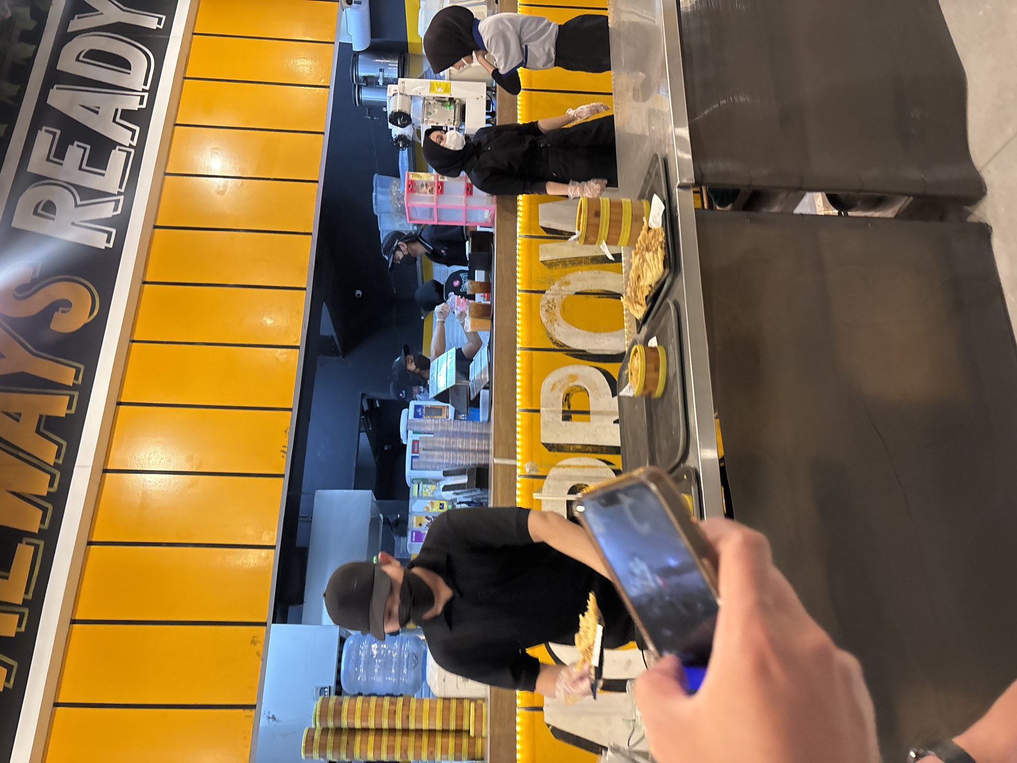 The image shows the interior of a restaurant, likely a fast-casual establishment, with a bright yellow wall adorned with text that reads "ALWAYS READY." In the foreground, a person's hand is visible holding a smartphone, angled towards a counter. On the counter, a plate of food, possibly fries or fried snacks, is presented in a black tray alongside a yellow cylindrical container.
Behind the counter, several staff members are engaged in their work. One staff member, wearing a black polo shirt and a black cap, is serving food. Another staff member, wearing a black hijab and a black uniform, stands near the serving area, while a third staff member, in a light blue and grey uniform and a hijab, is speaking to her. In the background, more staff members are visible, and a drink dispenser is present. The overall environment appears clean and organized, with stacks of yellow containers and a water cooler visible to the left. The context suggests this scene is likely within the city of South Tangerang, Indonesia, a common location for such eateries.