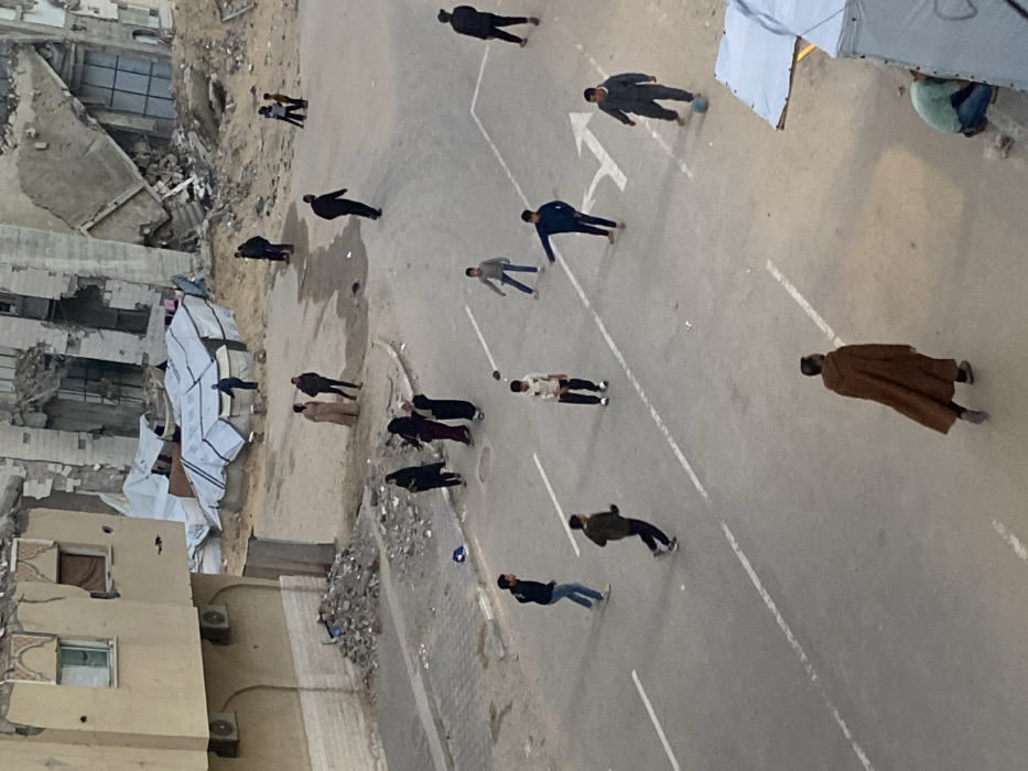 The image provides an aerial, slightly rotated perspective of a street scene in what appears to be a damaged urban or semi-urban environment. The overall impression is that of an area significantly affected by conflict or disaster, yet with individuals engaging in daily activities.
On the left side of the image, multiple multi-story buildings are severely damaged, with large sections reduced to rubble, exposed interiors, and collapsed walls. Piles of debris and dirt are visible around their foundations. Adjacent to these damaged structures, two large, light-colored tarpaulin sheets are draped over makeshift frames, forming temporary shelters or covers. Further to the left, a more intact building with a tan facade, several windows, and air conditioning units is visible.
The central and right portions of the image are dominated by a wide, paved street or open asphalt area. White painted markings are visible on the asphalt, including dashed lines that likely delineate lanes or parking spaces, and two large directional arrows. One arrow points towards the upper left (as oriented in the image), and the other points horizontally to the left.
Numerous individuals, predominantly appearing to be adult males, are scattered across the street. There are at least 15 to 20 people visible. Many are dressed in dark jackets, pants, and some wear more traditional long garments. One individual in the lower right stands out, wearing a long, loose brown robe, possibly a thobe or dishdasha.
A notable activity taking place is a casual game, likely soccer, as a small, light-colored ball is visible on the street. Several individuals are gathered around the ball or moving in its vicinity, suggesting active play. Other people are standing around, observing, or walking casually. Another smaller, blue spherical object, possibly a second ball or debris, is visible closer to the damaged buildings on the left side of the street.
The lighting in the image suggests daytime, possibly late afternoon or an overcast day, given the soft shadows and overall subdued light. The ground appears dry and dusty. The scene presents a stark contrast between the significant structural damage in the background and the ordinary human activity occurring on the street. No identifiable text, other than the street markings, is visible in the image.