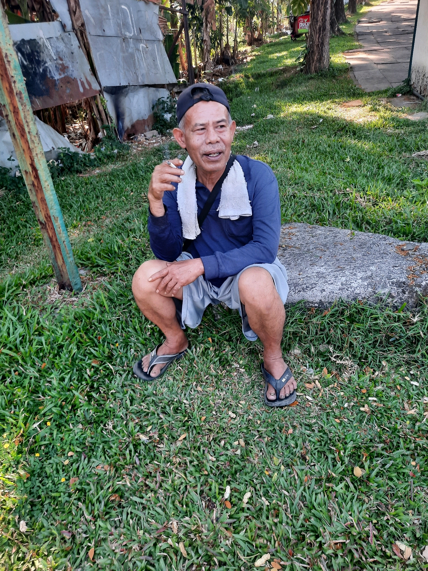 An older Asian man wearing a dark blue long-sleeved shirt, light gray shorts, a dark baseball cap, and dark flip-flops is seated on a large stone in a grassy area. He has a white towel draped around his neck and is holding a lit cigarette in his right hand. He appears to be exhaling smoke and looking to his right. In the background, there are trees, a pathway, and a makeshift structure constructed from corrugated metal. The scene is set in Babakan Madang, Indonesia, with the environment indicating an outdoor urban setting.