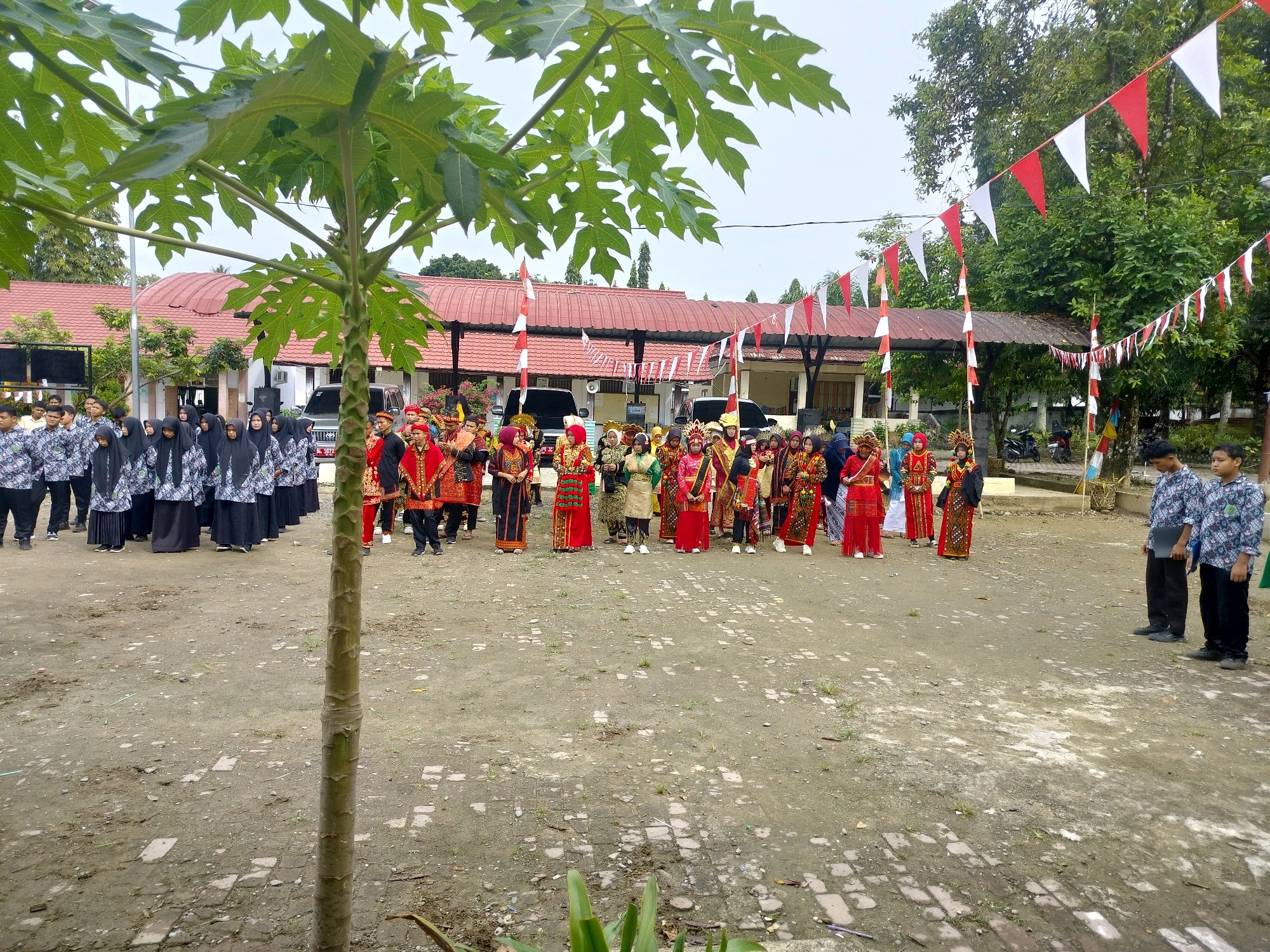 In a dusty outdoor area in Purwodadi, Indonesia, a group of approximately 40 people, including children and adults, are gathered. Many are dressed in colorful, traditional attire, suggesting a cultural event or performance. Some individuals are wearing black headscarves and traditional patterned shirts, while others are in brightly colored robes and elaborate headdresses. A line of red and white triangular flags strung across poles decorates the background, indicating a festive occasion. Two men in the foreground, dressed in patterned shirts and black pants, stand apart from the larger group. Behind them, a building with a red roof and awnings suggests a school or community center. The ground is a mixture of dirt and exposed brickwork. A papaya tree with large leaves stands prominently in the foreground, partially obscuring the view of some individuals. The overall atmosphere appears to be one of a community gathering or celebration.