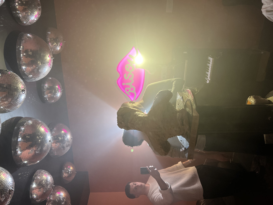 The image depicts an indoor scene within a venue identified as "Bisou" in Paris, France. The room features pink-hued walls and a dark ceiling from which numerous silver disco balls are suspended. Centrally positioned, an unglazed clay bust of a human torso, rough in texture and brownish in color, is displayed on a dark, cylindrical pedestal. To the left, a young male in a light-colored, long-sleeved sweater and dark trousers holds a dark smartphone upright, actively recording or photographing the scene. Behind the sculpture, two other individuals are partially visible; one male figure is largely obscured, while a female figure in a dark top is situated to the right, near a dark digital keyboard with prominent white keys. A strong, localized light source, likely a flash, is visible on the right, casting significant lens flare across the image. On the pink wall, a pink neon sign in the shape of lips, bearing the word "Bisou," is illuminated. The overall environment suggests an artistic event or performance taking place within a social establishment.