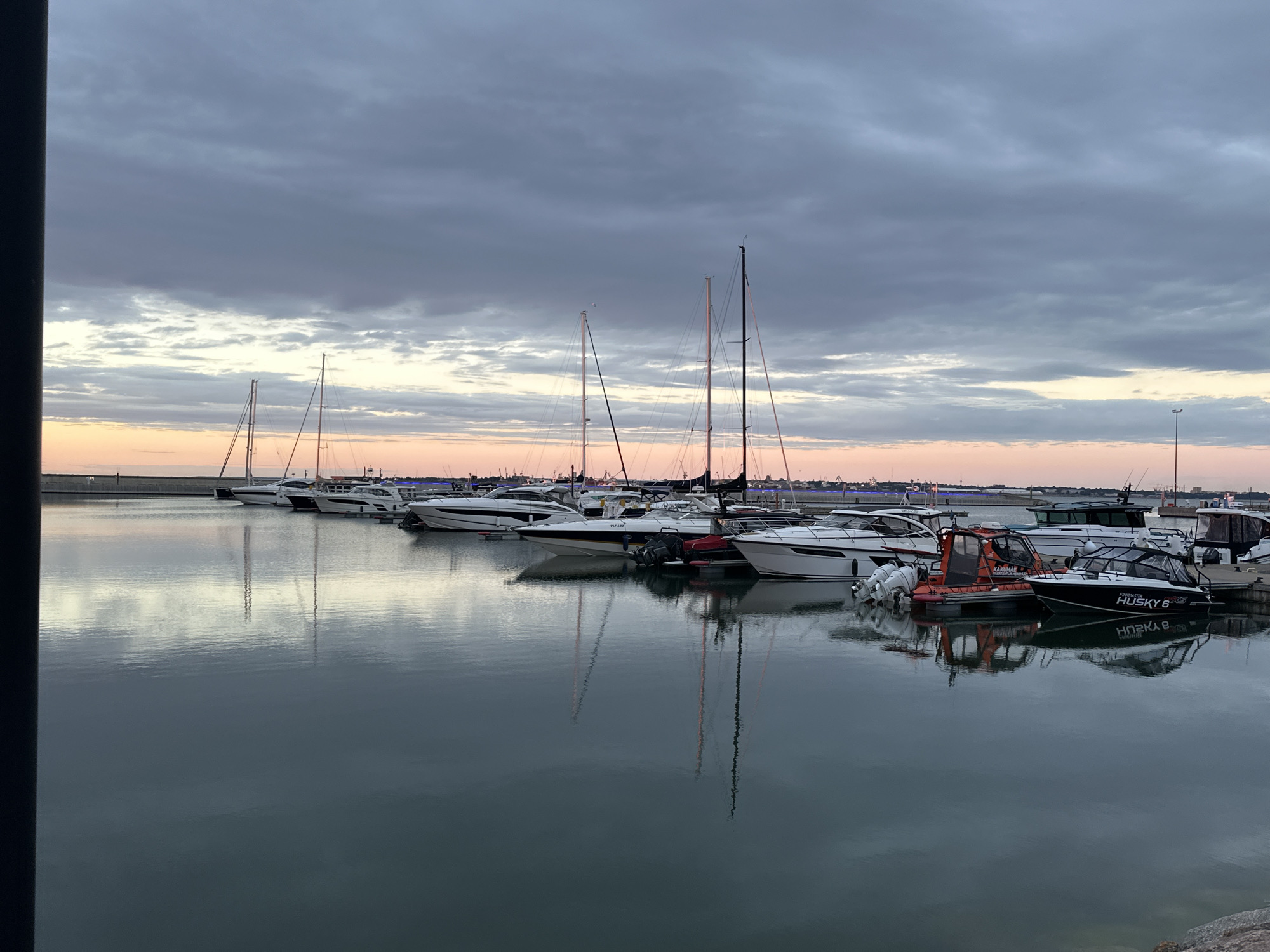 A harbor in Tallinn, Estonia, is depicted at dusk. Numerous boats, including yachts and smaller motorboats, are moored at docks. The sky is filled with dramatic, overcast clouds, tinged with the soft hues of sunset. The calm water of the harbor reflects the boats and the sky, creating a serene and picturesque scene.