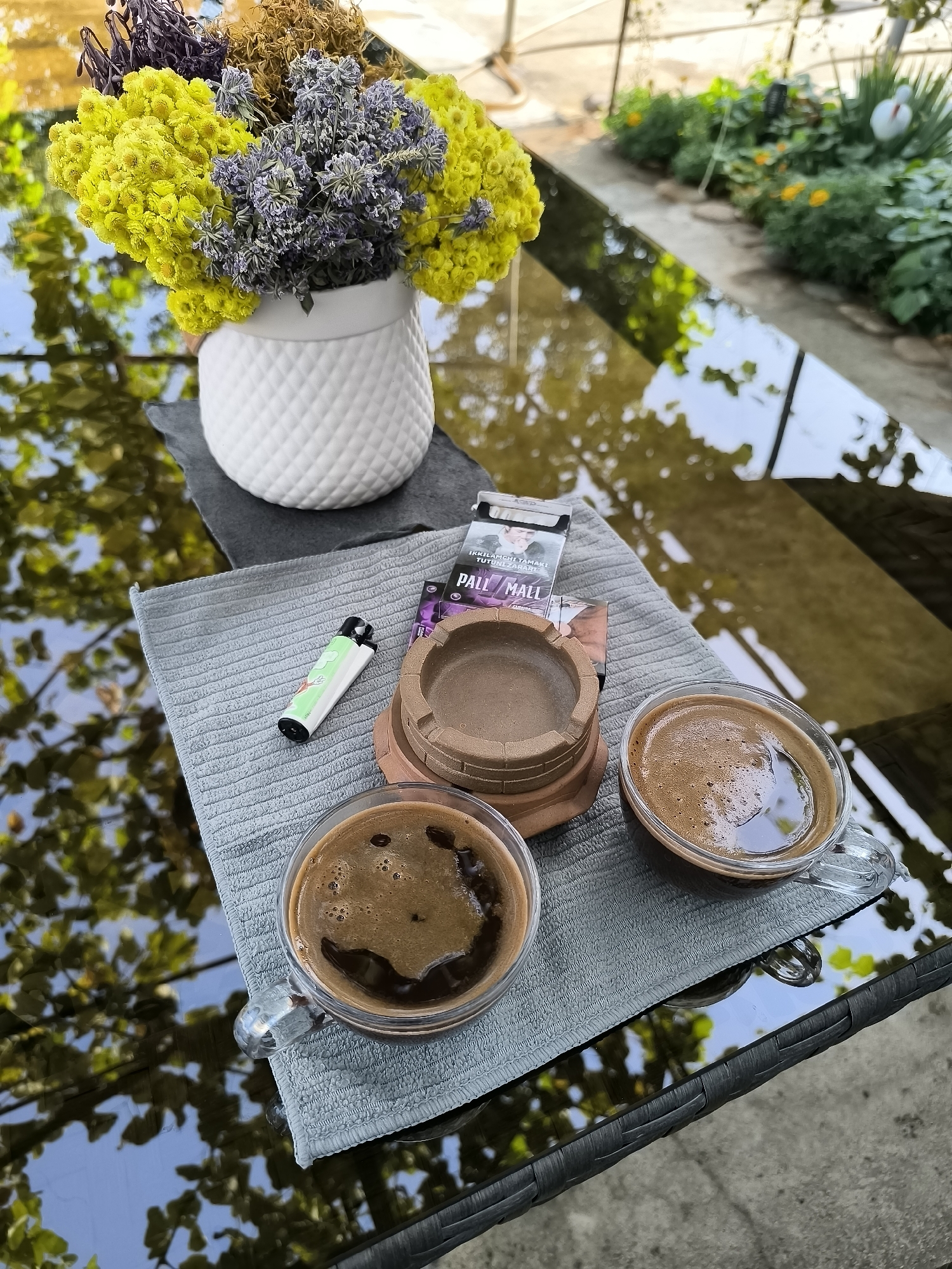This image shows a close-up view of two cups of coffee, a pack of Pall Mall cigarettes, a lighter, and a small ashtray placed on a grey fabric mat. The items are arranged on a reflective dark surface, possibly a glass table, which shows reflections of green foliage and a bright sky. In the background, a white ceramic vase filled with dried yellow and purple flowers is visible. Beyond the table, a garden area with lush green plants and yellow flowers can be seen, with a hint of a covered outdoor seating area. The overall scene suggests a relaxed outdoor setting, possibly in a cafe or a home garden. The location is identified as Almalyk, Uzbekistan.