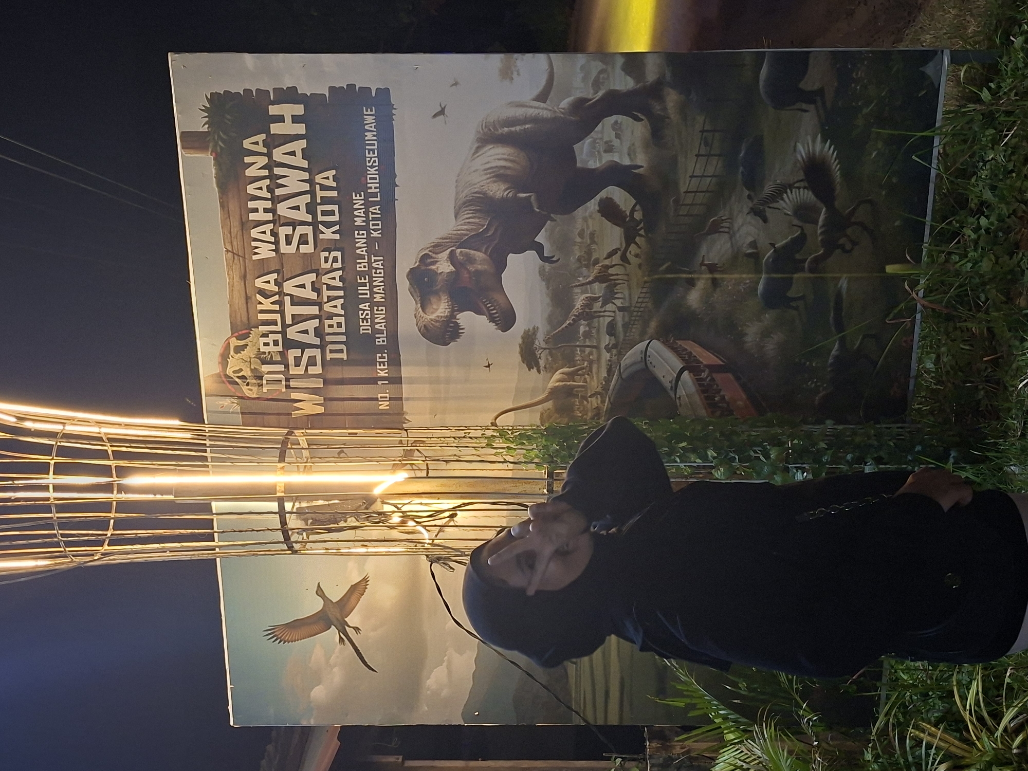 Here is a description of the image:
A woman wearing a headscarf is posing in front of a sign that reads "DIBUKA WAHANA WISATA SAWAH DIBATAS KOTA". The sign is a promotional board for a tourist attraction, featuring images of dinosaurs. The address of the attraction is indicated as "DESA ULE BLANG MANE NO. 1 KEC. BLANG MANGAT - KOTA LHOKSEUMAWE". Some light decorations are visible behind the sign and the woman. Green plants surround the scene.
