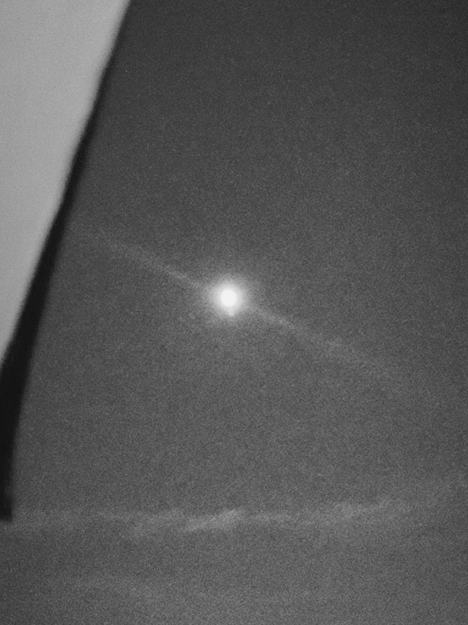 The image appears to be a monochrome photograph of a sky at night with a bright light source, likely the moon. Wispy clouds are visible near the bottom of the frame. In the upper left corner, there is a dark, undefined shape obstructing the view. The overall image has a grainy texture, consistent with low-light photography. There is no data present to ascertain the location in Nowy Tomyśl, Poland. No people, subjects, objects, actions, or interactions are visible other than the elements described above.