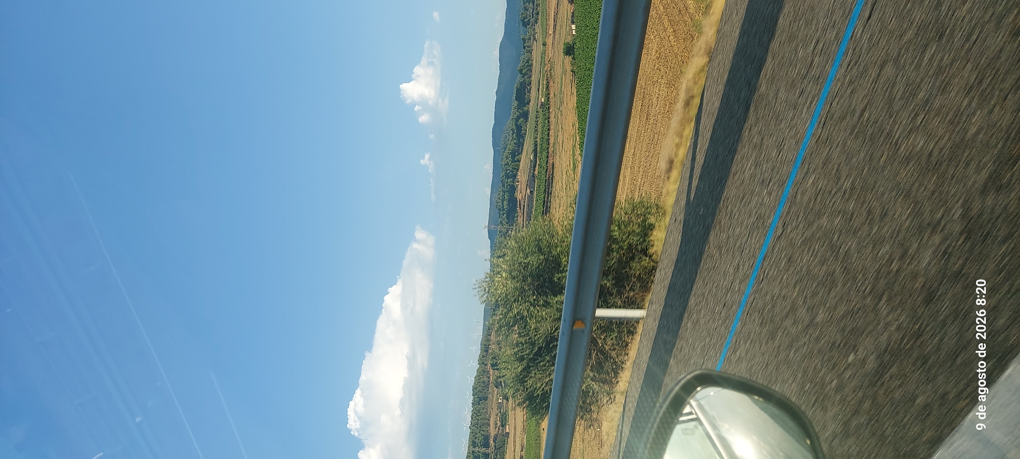 The image depicts a landscape scene with a clear blue sky and scattered clouds. In the foreground, a car's side mirror is visible on the left, reflecting the sky and some of the passing scenery. The road surface, with a distinct blue line, is in motion blur, indicating travel. A metal guardrail runs horizontally across the midground, separating the road from a field of dry, golden crops and patches of green cultivation. Further in the distance, rolling hills are covered with trees and dotted with a few buildings. Several wind turbines are visible on the distant hills. The timestamp "9 de agosto de 2026 8:20" is present in the bottom left corner, suggesting the image was taken on August 9, 2026, at 8:20 AM. The location is specified as L'Espluga de Francolí, Spain. The scene suggests a rural or agricultural area during daytime.
