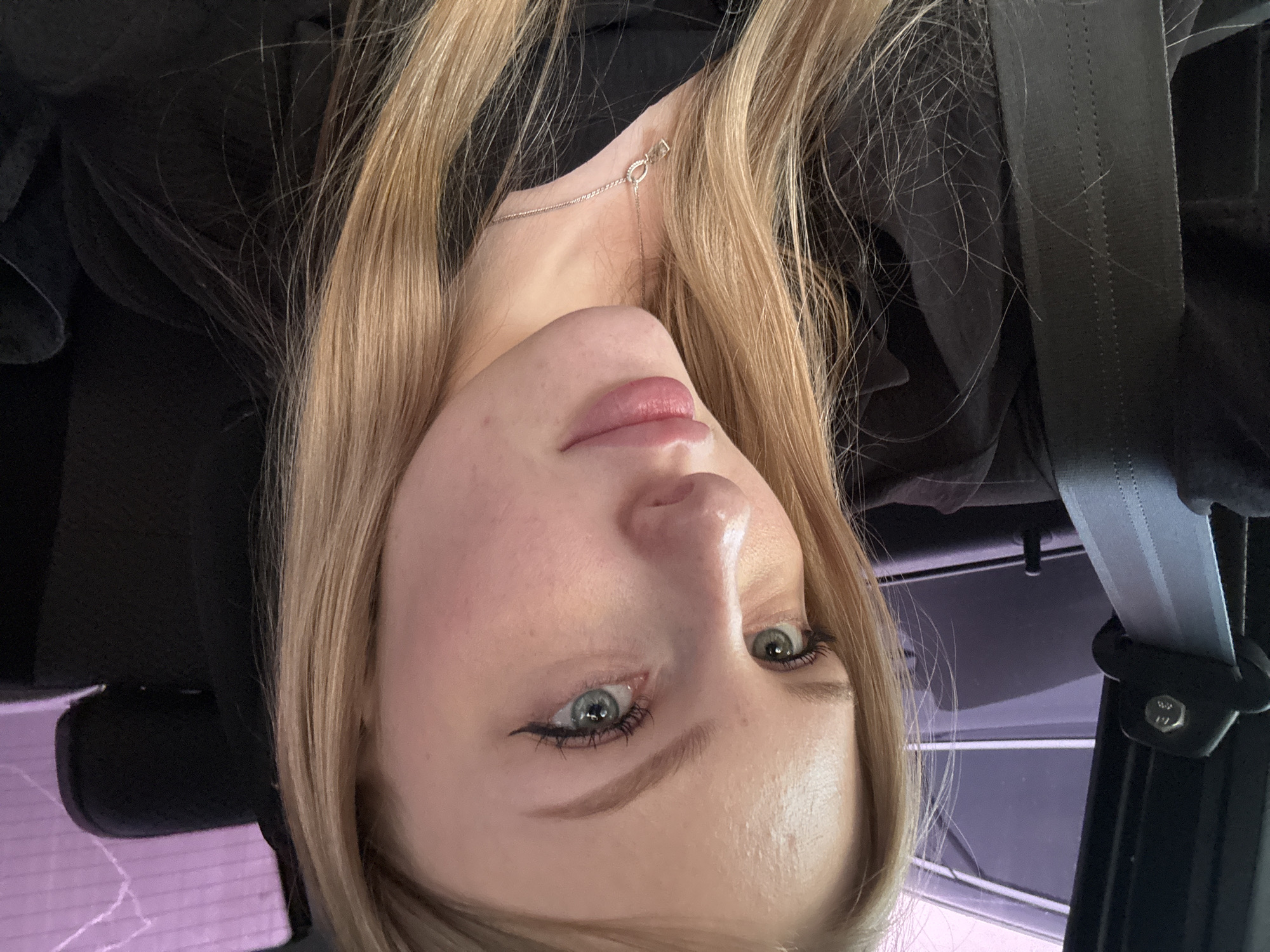 The image shows a young woman with blonde hair, blue eyes, and light skin. She is wearing a black top and a silver necklace. She appears to be in a car, as a seatbelt is visible. The background includes parts of the car interior, possibly a window with a lilac tint, and other unidentifiable car parts. It is not possible to determine if this image was taken in Кондратово, Russia from the provided information.