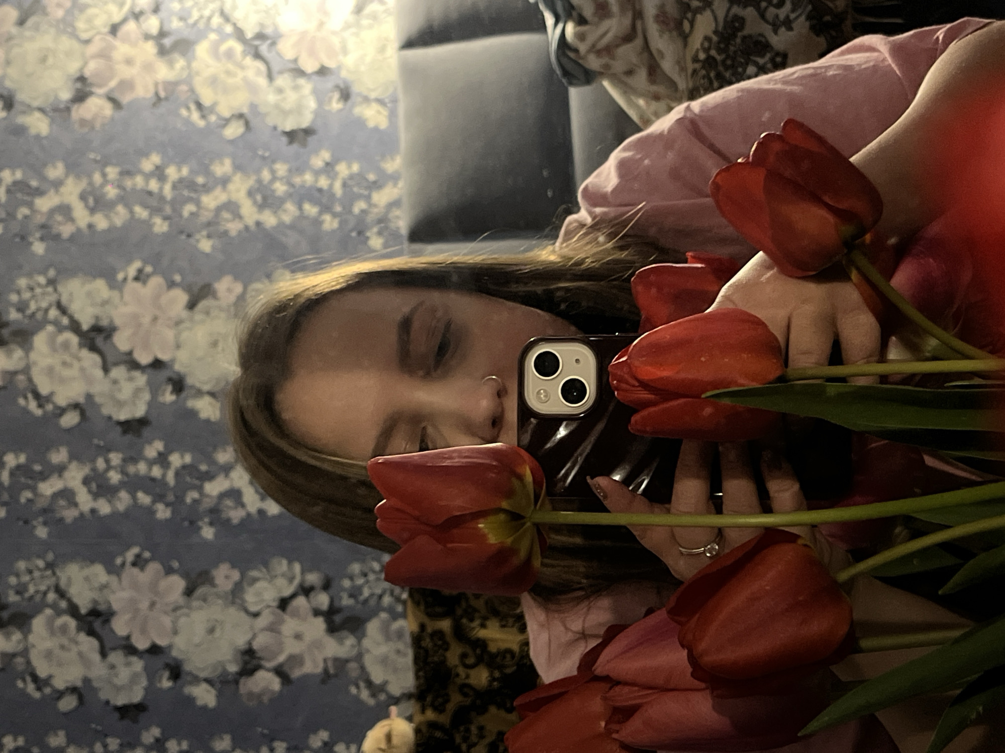 The image, presented with a 90-degree clockwise rotation, is a mirror selfie featuring a person holding a large bouquet of red tulips. The individual, appearing to be female, has long, dark hair and is wearing a light pink top. Her eyes are closed or cast downwards, and a small nose ring (septum piercing) is visible.
She holds a black smartphone, identifiable as an iPhone due to its dual camera system, in her left hand to capture the reflection. Her right hand, adorned with a ring featuring a clear stone and manicured nails, holds the stems of the vibrant red tulips. The tulips themselves are a prominent feature, with their deep red petals and some green stems visible throughout the bouquet.
Reflected in the mirror behind the person is a room interior. The background wall displays blue-grey wallpaper patterned with subtle, light-colored floral motifs. A dark piece of furniture, possibly a sofa or headboard, is also visible in the upper background. The lighting in the scene appears to be soft and ambient, suggesting an indoor setting. There is no visible text in the image.