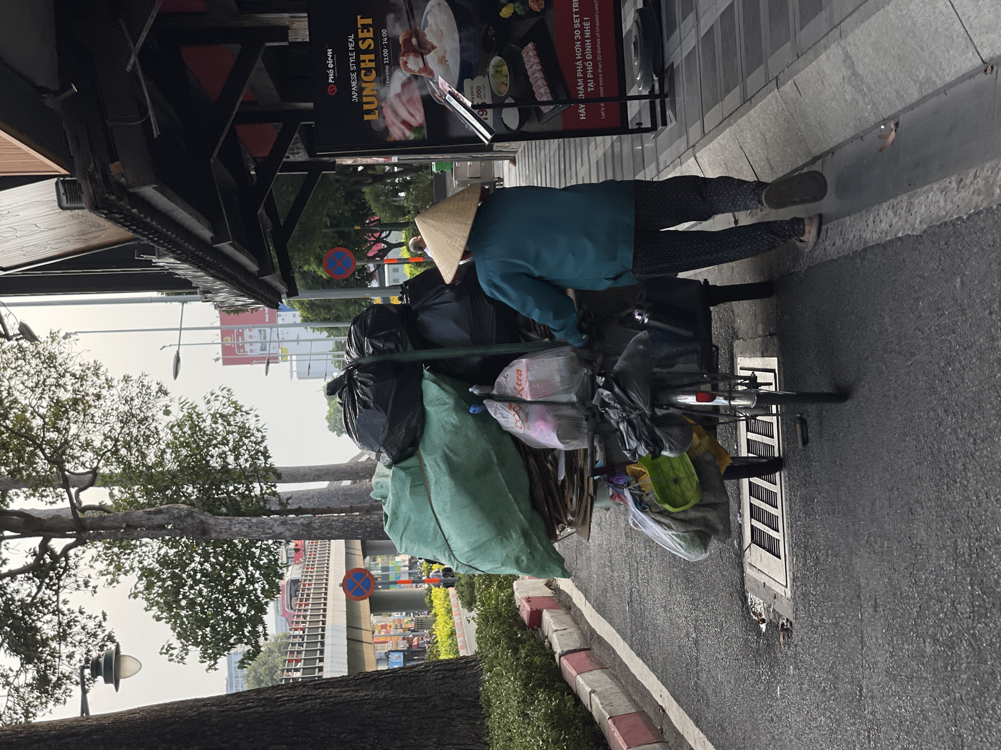 A person wearing a conical hat and a blue jacket stands next to a bicycle heavily loaded with bags, likely collecting recyclables in Thủ Đức, Vietnam. The bike is parked next to a sidewalk with a restaurant advertisement board behind the individual. The background includes trees, a street, and elevated infrastructure.