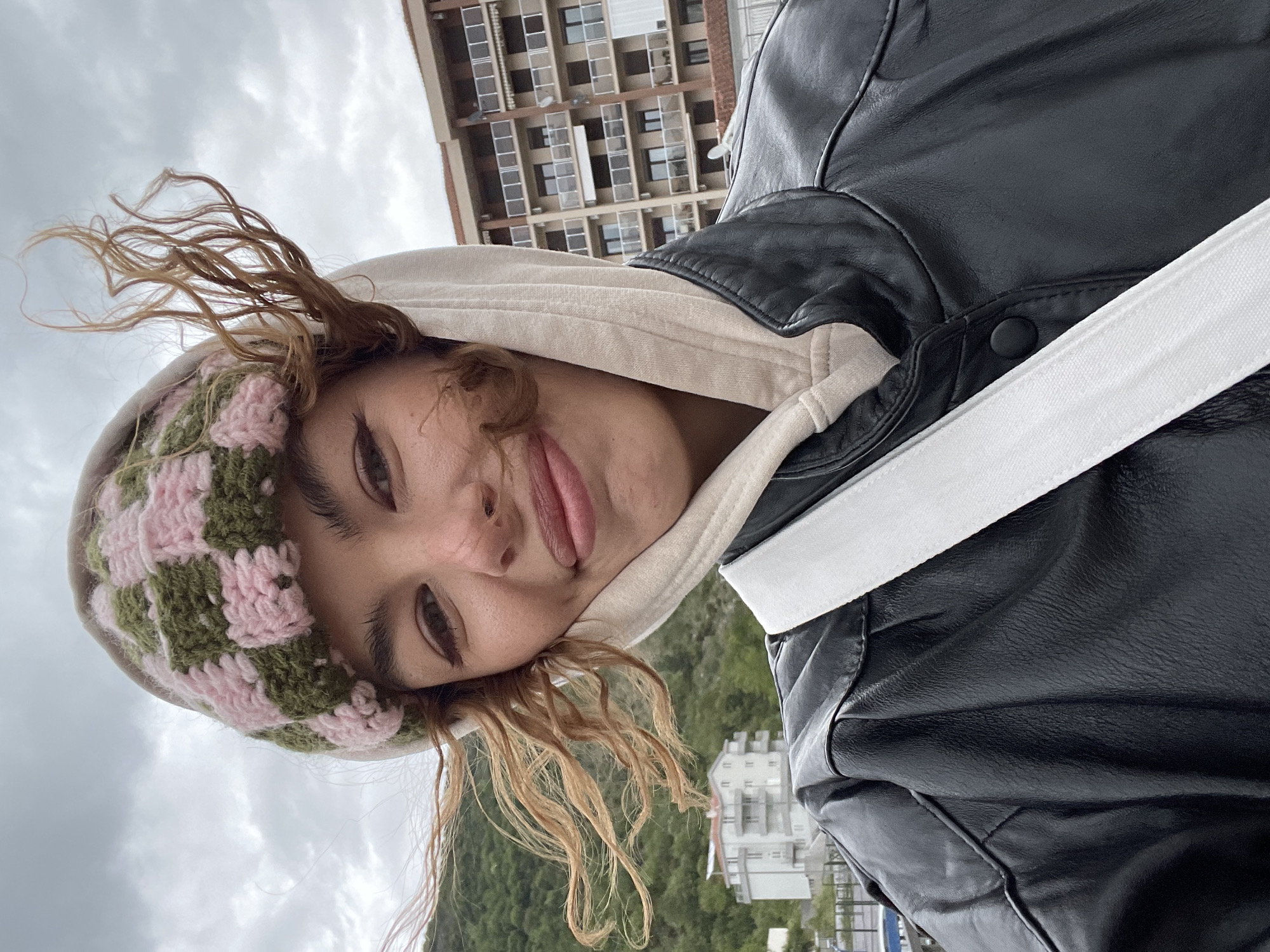 A casual outdoor selfie captures a young person, likely female, with their head tilted significantly to the left. They look directly at the camera with a relaxed, playful expression, a subtle smile, and their tongue slightly visible between their lips. Their light brown, wavy hair appears windswept, fanning out around their face.
The person wears a knitted beanie with a checkerboard pattern in pink and olive green. Beneath, a beige hooded sweatshirt is visible, topped with a black faux leather or leather jacket. A white strap, possibly from a shoulder bag, crosses diagonally across their chest.
The background shows an overcast, cloudy sky. In the distance, a tall, multi-story apartment building with numerous windows is visible. Lush green hills or mountains stretch across the mid-ground and background, with a smaller white building nestled amongst them. The scene suggests a daytime setting in an elevated location, possibly overlooking a coastal area, consistent with Boreti, Montenegro. There is no visible text in the image.