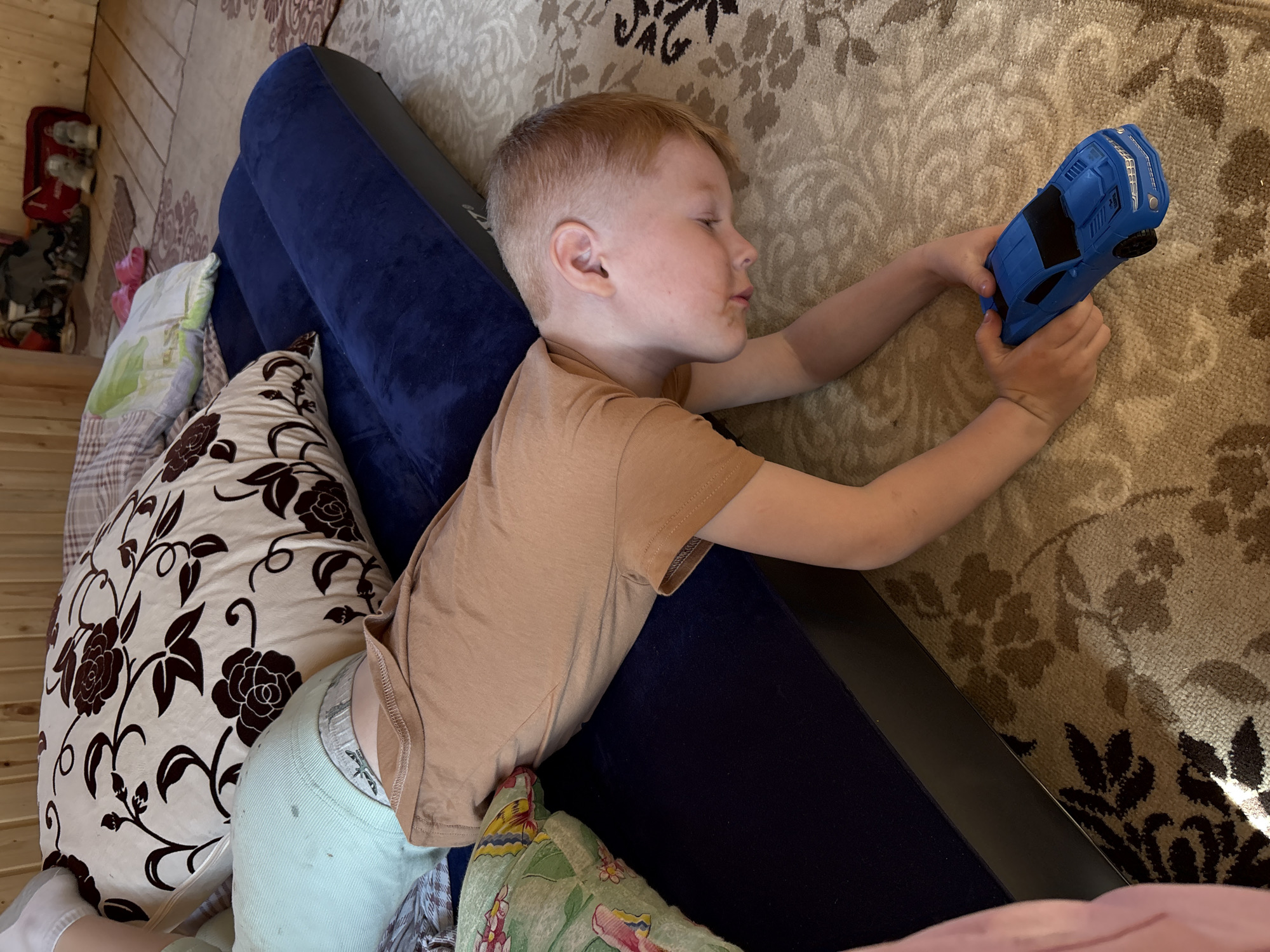 The image depicts an indoor scene in Italmas, Russia, featuring a young male child, estimated to be between 4 and 7 years old, with short, light-colored hair. The child is lying prone, with his upper torso and arms on a light-colored, patterned rug, and his lower torso and legs resting on a dark blue inflatable mattress. He is wearing a light brown short-sleeved t-shirt, with a gray patterned waistband of underwear visible above the t-shirt's hem. His head is turned to the right, facing down towards the rug, and both hands are holding a bright blue plastic toy car, which appears to be a sports car model.
The dark blue inflatable mattress, which has a plush, possibly velvet flocked surface, is positioned on the rug. Behind the child's lower body, on the mattress, rests a large, cream-colored pillow adorned with a repeating dark brown floral pattern of roses and vines. Another lighter-colored pillow is partially visible beneath the patterned pillow. Further into the background, a smaller, light green pillow with a vibrant pink and yellow floral design is visible.
The floor is predominantly covered by a light beige rug with a repeating pattern of dark brown and tan floral and vine motifs. The background reveals light-colored wooden plank walls, suggesting a rustic interior. On a visible section of wooden floor in the far background, several pairs of shoes, including a pair of white furry boots, and a red duffel bag are present.