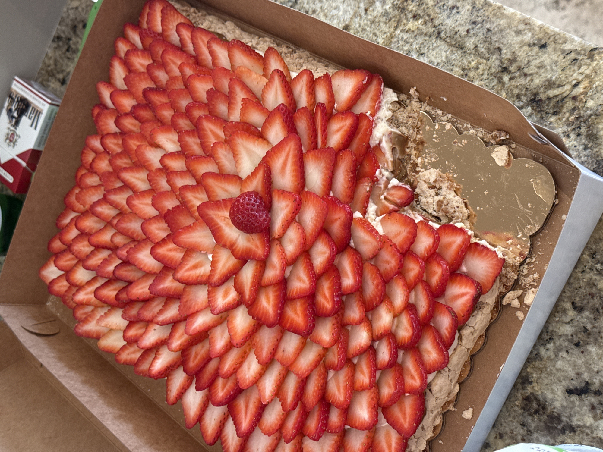 The scene depicts a partially consumed rectangular dessert presented in an open brown cardboard box on a speckled granite-style countertop in Miami Beach, United States of America. The dessert is a multi-layered cake or tart, generously topped with an arrangement of uniformly sliced, bright red strawberries laid in overlapping rows. A single, small, whole strawberry is positioned centrally among the sliced fruit. A creamy white filling is visible beneath the strawberry layer. The base of the dessert consists of a light brown, crumbly, and textured pastry, likely meringue. A portion of the dessert, approximately one-quarter, has been removed from the bottom right, revealing the internal layers and a golden scalloped cardboard cake board beneath. Crumbles of the pastry base are scattered inside the box around the missing section. In the upper right background, a red and white Marlboro cigarette pack rests on the countertop, next to a partially visible green object. The cardboard box has cut-out handle features on its sides and a silver-colored edge along its base.