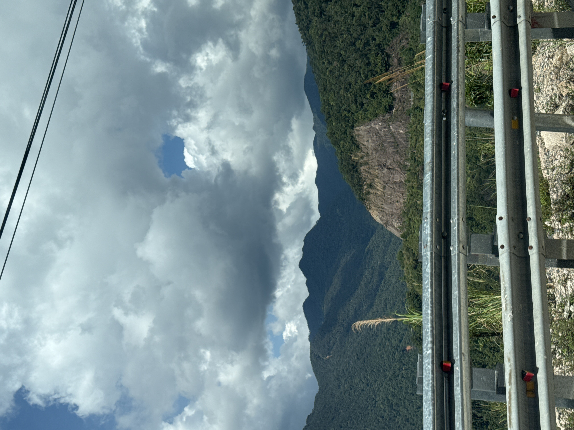 An eye-level, medium shot captures a scenic mountain landscape under a cloudy sky. In the foreground, a silver guardrail with reflective red and yellow markers runs horizontally across the frame. Behind the guardrail, tall, dry grass and some green vegetation are visible. The background features a dramatic mountain range covered in dense green foliage. A large, rocky cliff face is prominent on the right side of the mountains. The sky is filled with dramatic, grey clouds, with a small patch of blue visible in the upper center. Overhead, two dark power lines stretch across the top of the image. The scene appears to be captured from a moving vehicle on a road, likely in Nam Khanh Vinh, Vietnam, given the lush mountainous terrain and the presence of a guardrail common on roads in such regions. No people are present in the image.