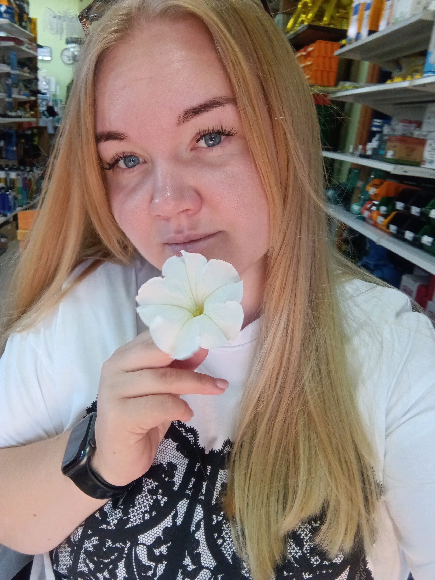 A medium shot shows a young woman with blonde hair and blue eyes holding a white flower near her face. She is wearing a white t-shirt with a black lace-like pattern on the front. A black smartwatch is visible on her left wrist. The background appears to be a retail store with shelves stocked with various items, including bottles and boxes. The lighting is natural and suggests an indoor setting. The image was captured in the city of Fokino, Russia.
