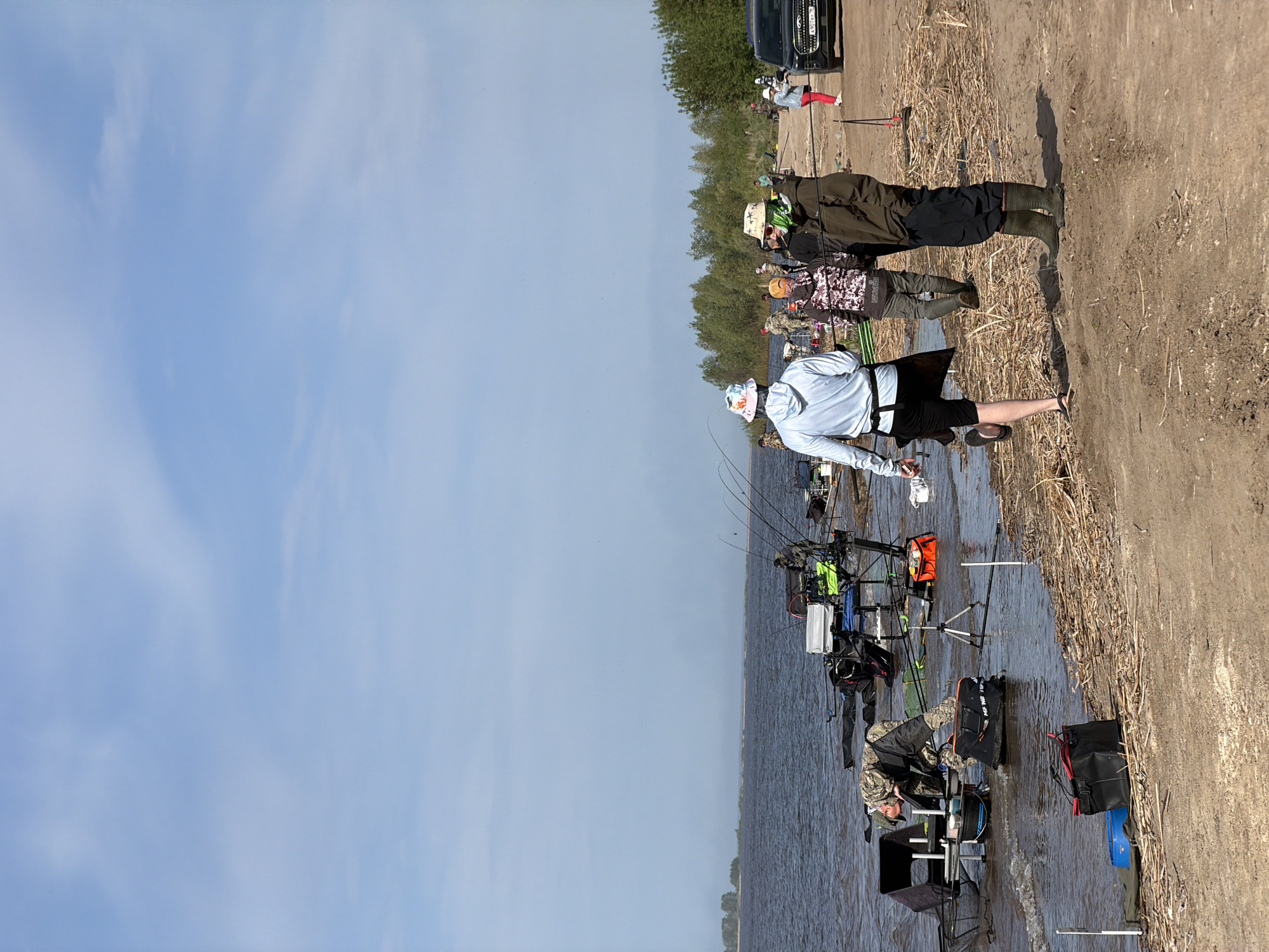 The image captures a fishing competition taking place on the shore of a body of water, likely in Krasnyi Bor, Russia. Multiple individuals are visible, equipped with fishing rods and gear, stationed along the shoreline. The setting includes a sandy bank transitioning into shallow water, where anglers are positioned. Fishing equipment such as rods, stands, and various containers are scattered around each person, indicating organized activity. The sky is clear and indicates daytime.