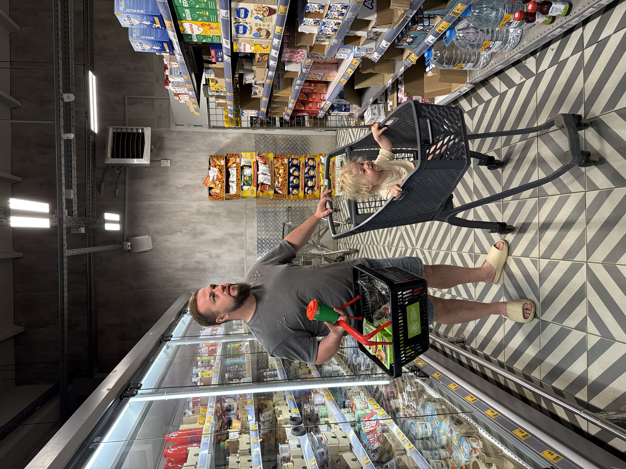 The image captures a detailed scene within a supermarket aisle in Sobinka, Russia. An adult male and a toddler are positioned in the foreground, engaged in grocery shopping.
The adult male, exhibiting a beard and short light-brown hair, is clad in a grey, distressed t-shirt, denim shorts, and light beige slip-on sandals. He stands with his left hand on the handle of a dark grey shopping cart, and his right hand holds a small black plastic shopping basket containing dark-wrapped items. A green cup with a red lid is also held in his right hand, positioned above the basket. His gaze is directed towards his right, past the refrigerated display.
A toddler, with curly blonde hair and wearing a light-colored t-shirt, is seated inside the main compartment of the dark grey shopping cart, facing towards the viewer's right. The toddler appears to be smiling, with both hands resting on the edge of the cart.
To the left of the scene, a long refrigerated display case with glass doors extends along the aisle. It is stocked with various dairy products, including yogurts and cheeses, and features numerous yellow price tags. To the right, multiple metal shelves are stocked with a variety of packaged groceries. Notable products include baby formula (Nestogen, Nutrition, Nestle), cereals, and milk products. Many shelves exhibit gaps, indicating sold items. The lowest shelf displays large clear bottles of water and smaller red-capped bottles. In the background, center-left, a tall, stacked display of "Lay's" potato chips and sunflower seed packets is visible. Mounted on the back wall, center-right, is a white industrial fan or heater unit labeled "VOLCANO."
The supermarket features an industrial aesthetic with dark grey, textured concrete walls and a dark ceiling crisscrossed with metal conduits and exposed lighting fixtures. The floor is tiled with a distinctive chevron pattern in alternating shades of dark and light grey.