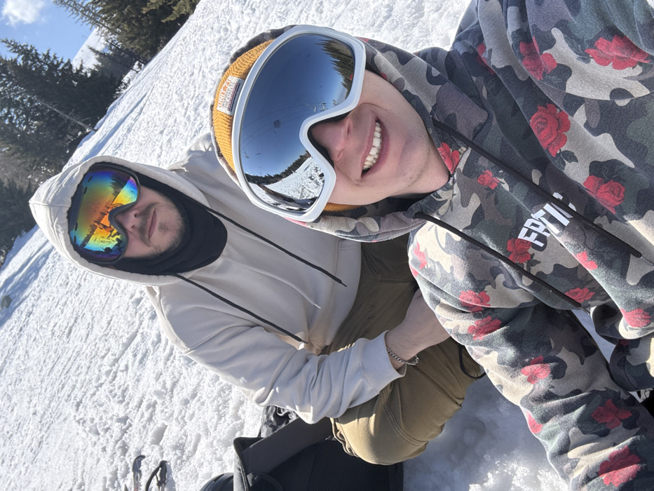 The image captures two male individuals positioned on a sunlit, snow-covered mountain slope, implied to be located in Bistritsa, Bulgaria. The foreground subject is a male, smiling and looking directly at the camera. He wears a camouflage-patterned hooded sweatshirt featuring red rose designs and a white textual print on the chest. An orange knitted beanie is visible beneath his hood, and he wears large white-framed ski goggles with mirrored lenses reflecting a ski lift and snowy terrain. Behind and slightly above him, a second male subject looks forward. This individual has facial hair, wears a light beige hooded sweatshirt with the hood up, a black neck gaiter covering his lower face, and large ski goggles with highly reflective, rainbow-colored lenses. He wears tan-colored pants and a silver chain bracelet on his left wrist. A black object, consistent with a backpack or gear bag, is partially visible on the snow to the left of the background subject. The environment features a steep, snow-covered incline with scattered evergreen trees in the mid-ground and a clear blue sky with light clouds above.
