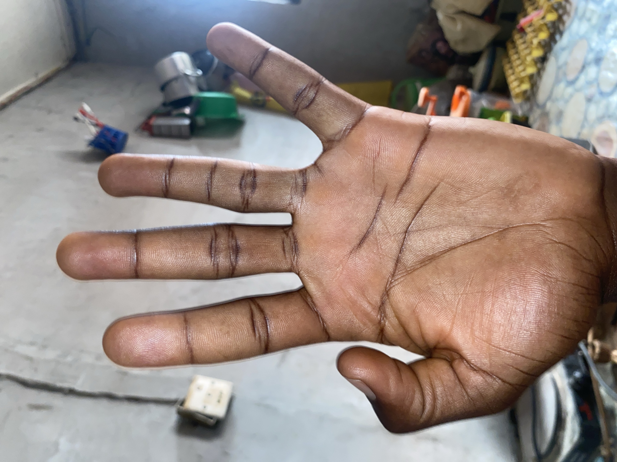 A close-up view of a dark-skinned human hand, palm facing the camera. The hand displays four extended fingers and a slightly bent thumb. The background appears to be a room or interior space with various objects scattered on a light-colored surface, including kitchenware, electrical components, and packaged goods. There is no clear indication of a location within Barden Yana, Nigeria. The environment seems to be indoors. There are no visible actions or interactions. The light is bright and somewhat diffused.