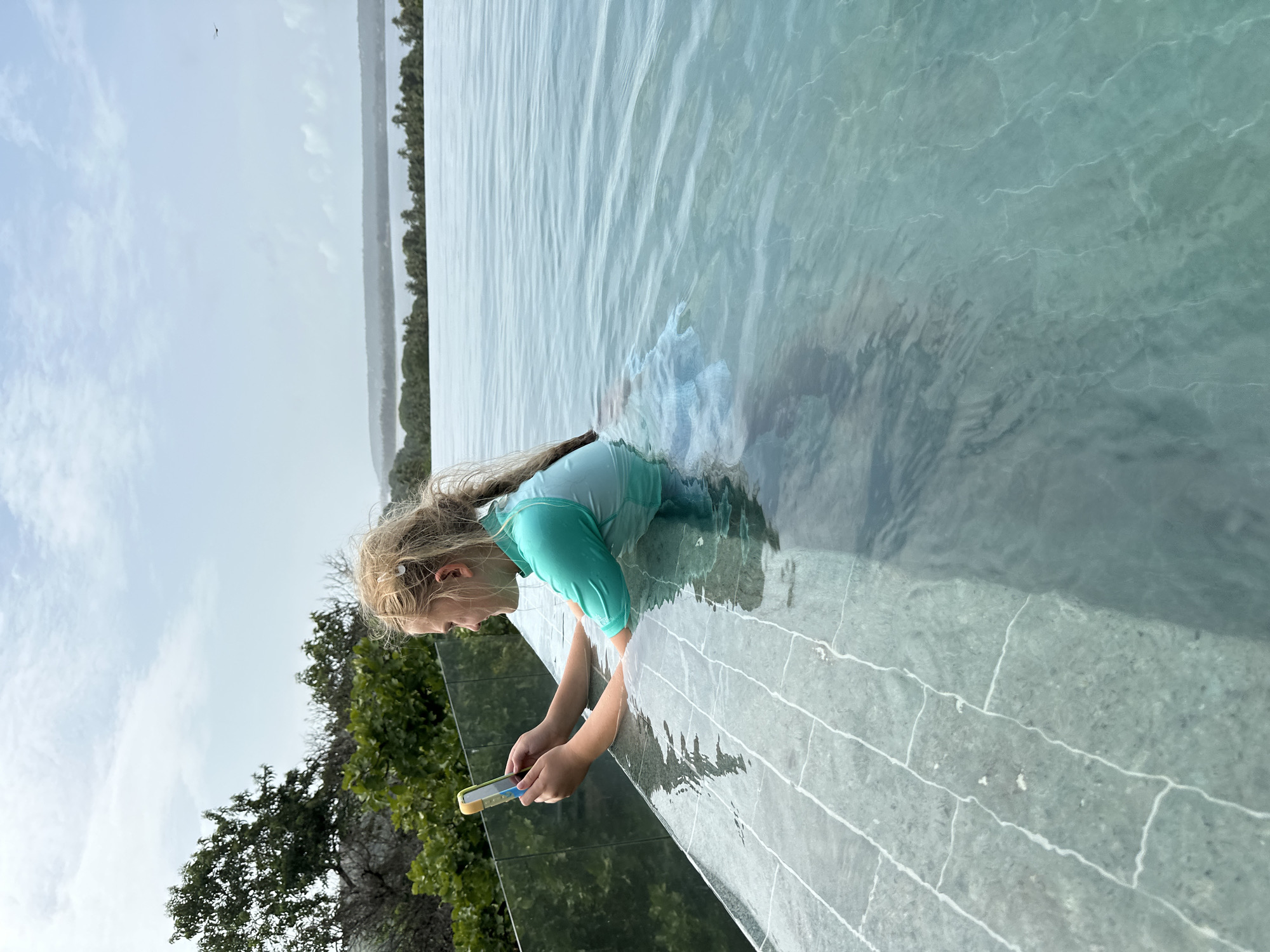 A young blonde girl in a turquoise rash guard is partially submerged in an infinity pool, leaning on the edge and holding a smartphone. She is looking at the screen of the phone. The pool has stone tiling on its edge, and the water is clear and blue. Beyond the pool, lush green trees and a body of water can be seen, suggesting a coastal or tropical location. The sky is partly cloudy. The scene appears to be in the city of Vagator, India, a known destination for resorts with infinity pools and ocean views.