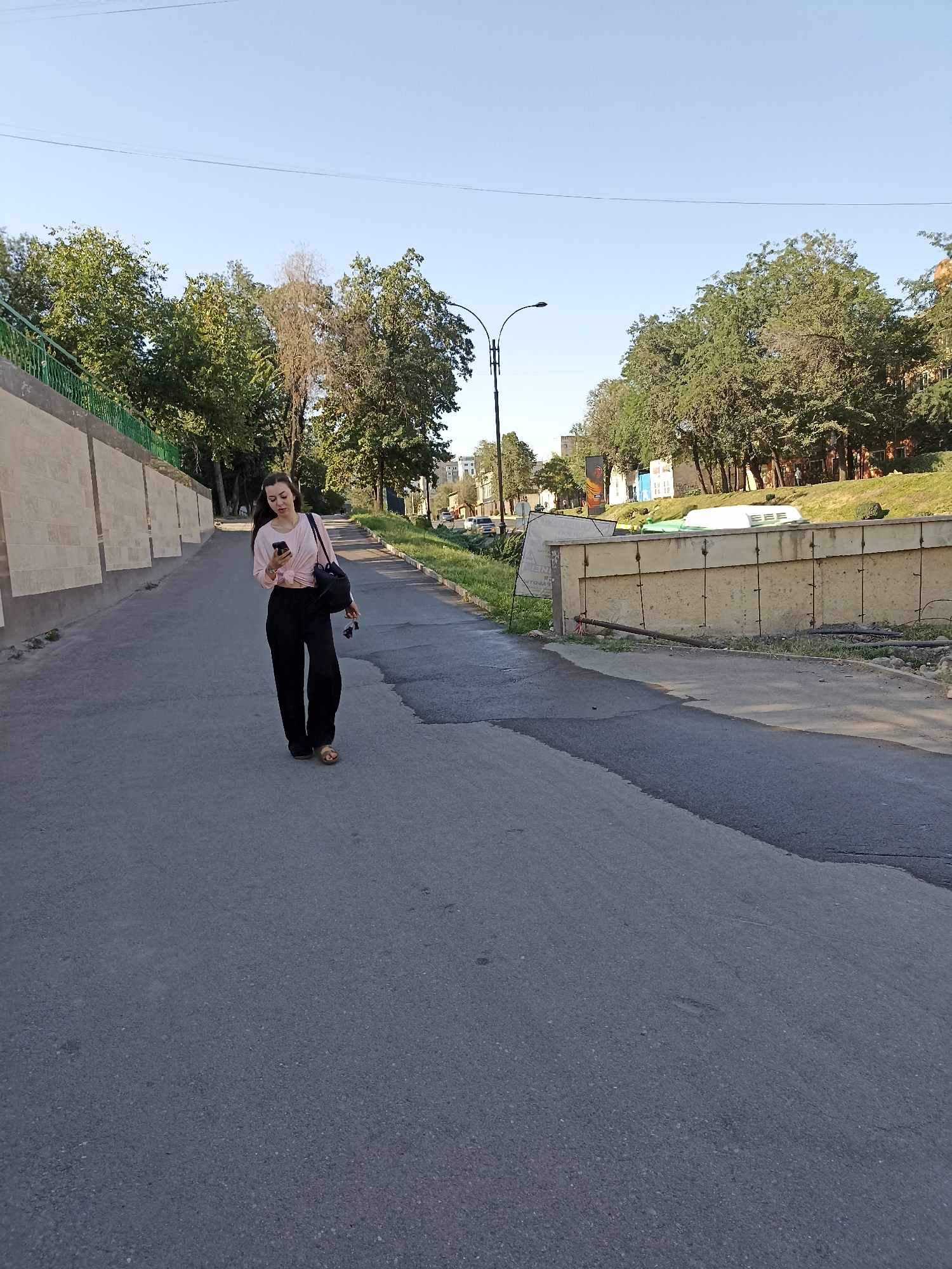 A young woman with long dark hair is walking down a paved pathway, looking at her smartphone. She is wearing a light pink, knot-front blouse and black, wide-leg pants, with brown sandals. A black backpack is slung over her left shoulder. The pathway is bordered on the left by a tan, concrete retaining wall with a green metal railing on top. On the right, the pathway is bordered by a grassy slope with trees and a concrete barrier. In the background, there are more trees, a street lamp, and some buildings. The scene appears to be in an urban park or a tree-lined street in Bishkek, Kyrgyzstan, during daylight hours under a clear sky.
