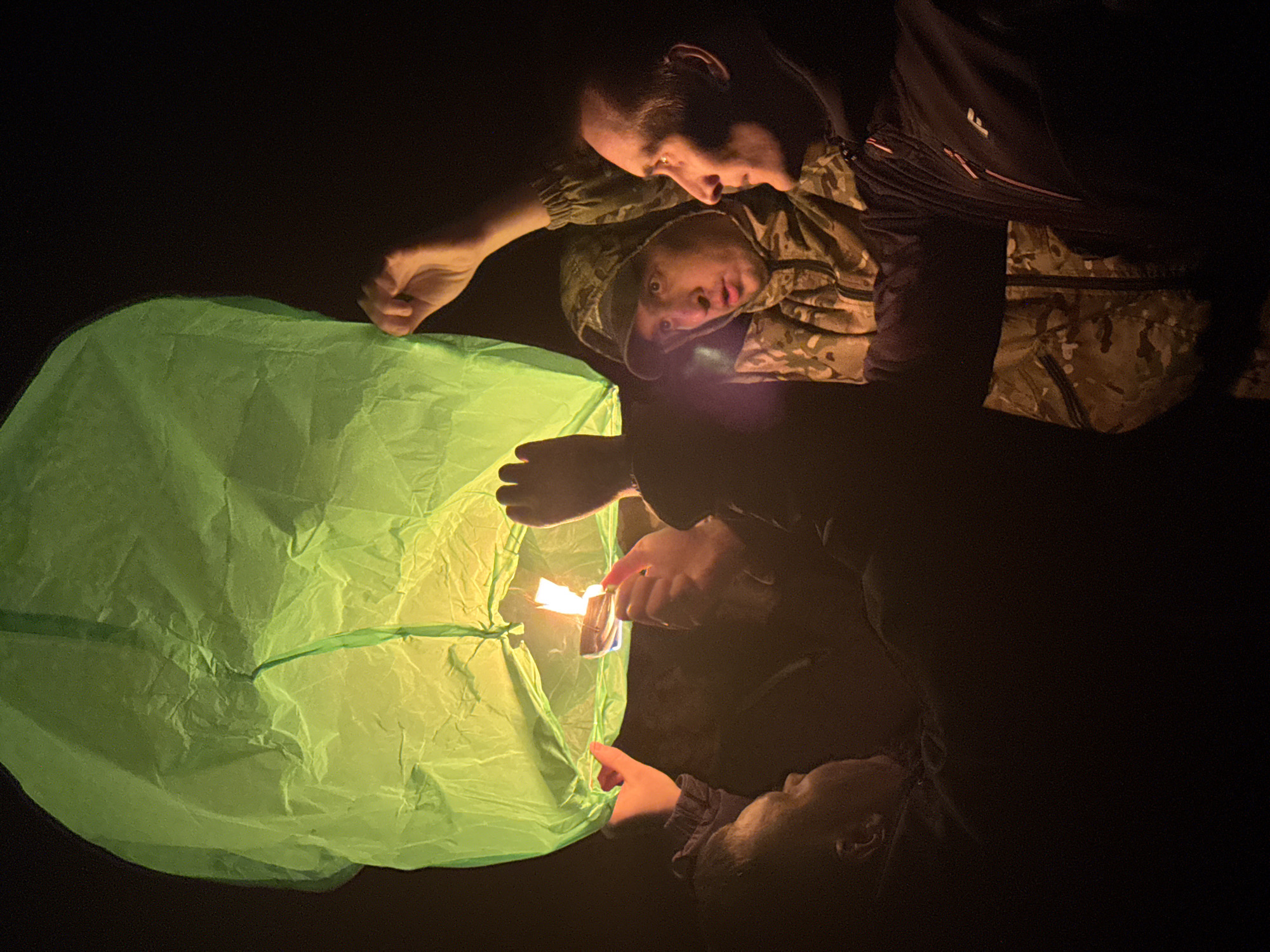 In a dark outdoor setting, an adult male and a child are jointly launching a green sky lantern. The adult, dressed in a black jacket with red accents over a camouflage patterned garment, holds the lantern open with one hand while using a lighter held in the other to ignite the fuel source at the base of the lantern. The child, wearing a camouflage hooded jacket, looks on with interest. The light from the burning fuel illuminates the lantern and the faces of the individuals. The scene is in Chaikovsky, Russia.