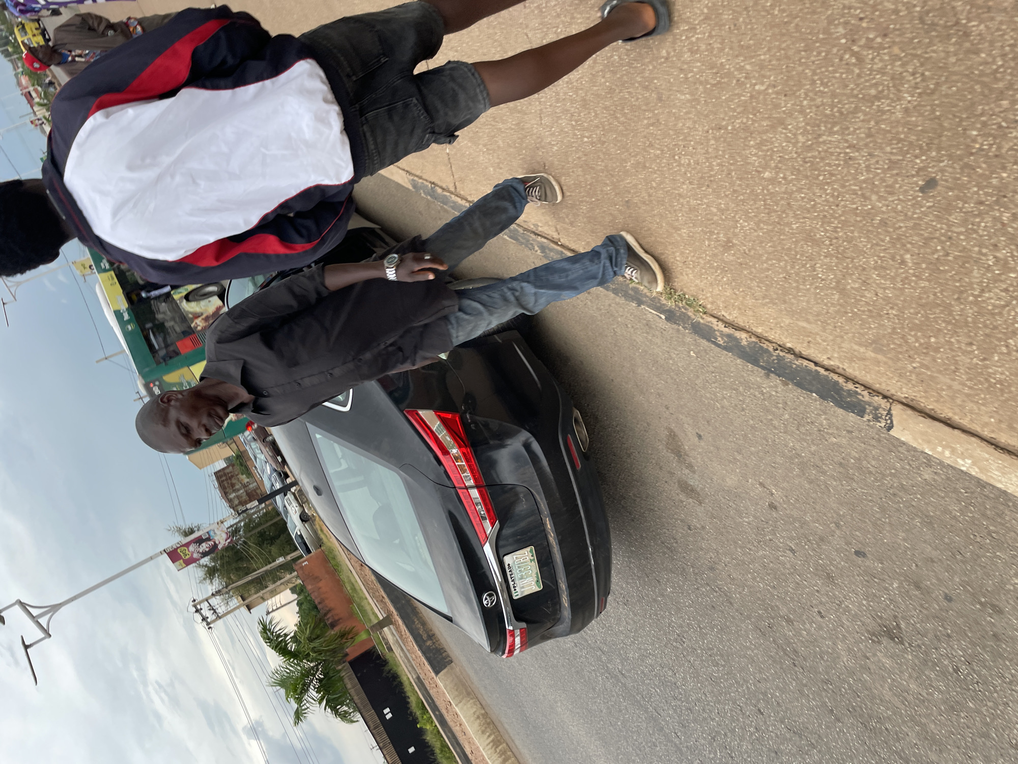 Two men are standing by a black Toyota sedan on a street in Jos, Nigeria. The man on the left, wearing a black button-down shirt and blue jeans, is looking down. He has a watch on his left wrist. The man on the right, wearing a white, red, and blue jacket and dark shorts, is facing away from the camera, partially obscuring the view of the car's rear. A yellow bus is visible in the background, along with buildings and streetlights. The sky is overcast. The car's license plate is visible, reading "JJN 337 BZ". The street is paved, with a curb and sidewalk. The scene appears to be a public area within the city.