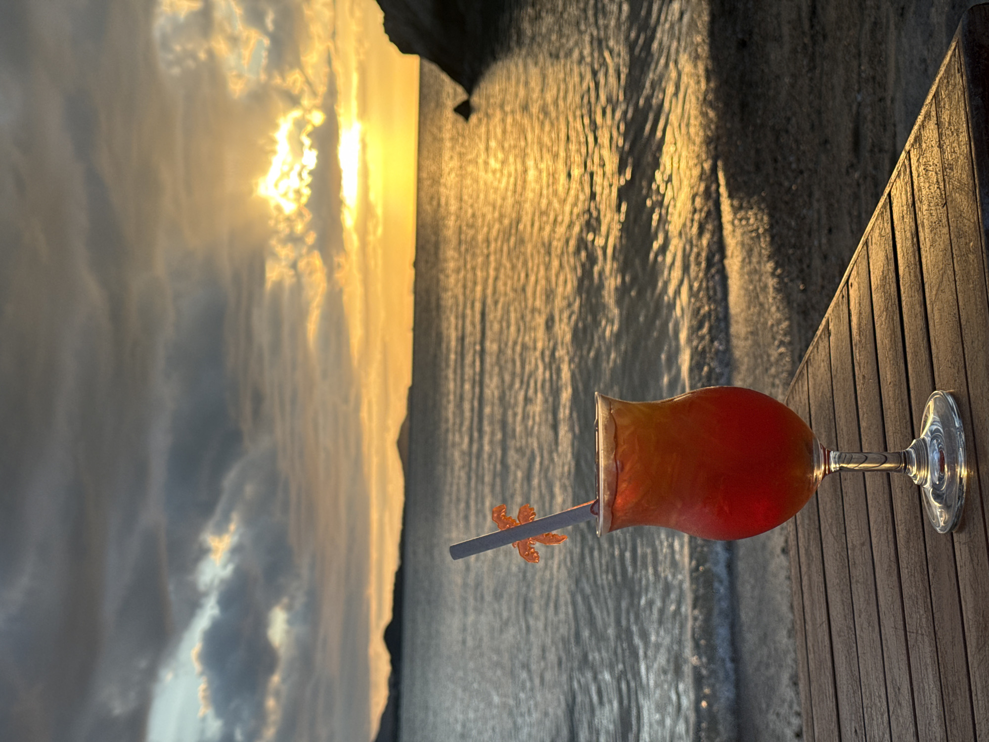 A colorful cocktail sits on a wooden deck overlooking the ocean at sunset. The drink is layered with red and orange hues, and a grey straw with an orange starfish garnish extends from it. The sun, low in the sky, casts a warm, golden glow across the water, creating shimmering reflections. Clouds in the sky are tinged with yellow and orange from the setting sun. The scene suggests a relaxing evening in a tropical location, likely a beach resort, given the presence of the wooden deck and the ocean. The overall mood is peaceful and serene. No text is visible in the image.