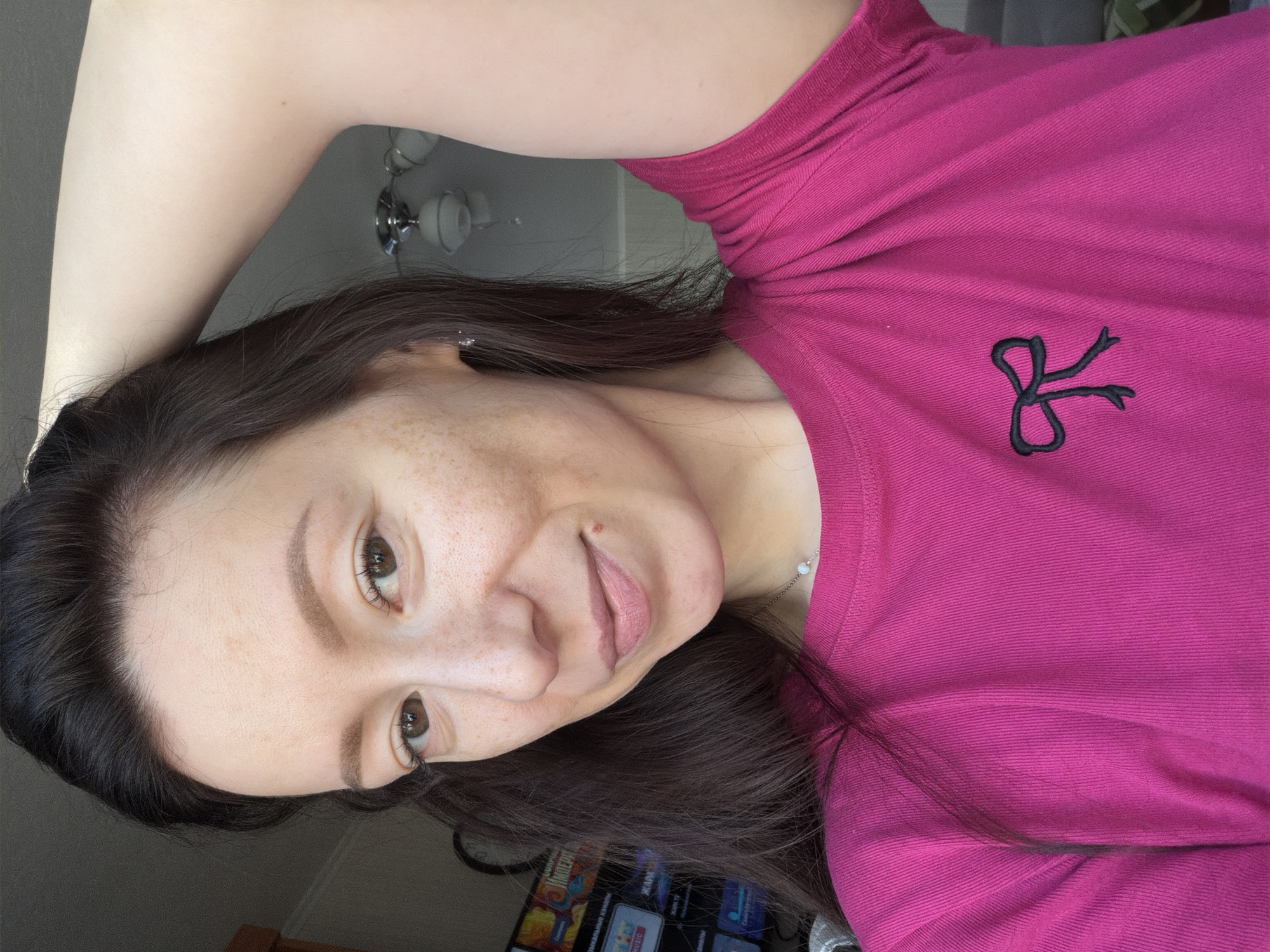 A woman with brown hair and freckles is wearing a pink t-shirt with a black logo. She is holding her left arm above her head. The background is a room with a ceiling light and a television. The location is Raevka, Russia.
