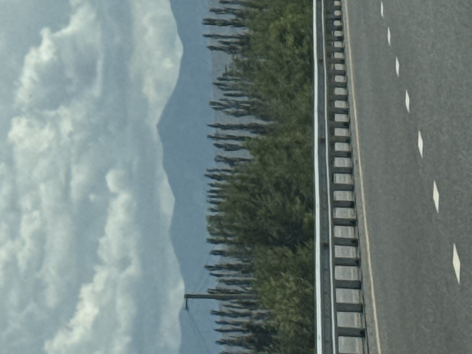 This image depicts a cloudy sky over a landscape that includes mountains in the distance, a row of tall trees, and a road with a guardrail. The foreground shows the asphalt road with white markings. A single utility pole is visible among the trees, and the trees themselves are dense and appear to be evergreen. The mountains are hazy blue in the background. This scene is consistent with the scenery found in and around the city of Balykchy, Kyrgyzstan, which is known for its location on the western shore of Issyk-Kul Lake and its mountainous surroundings.