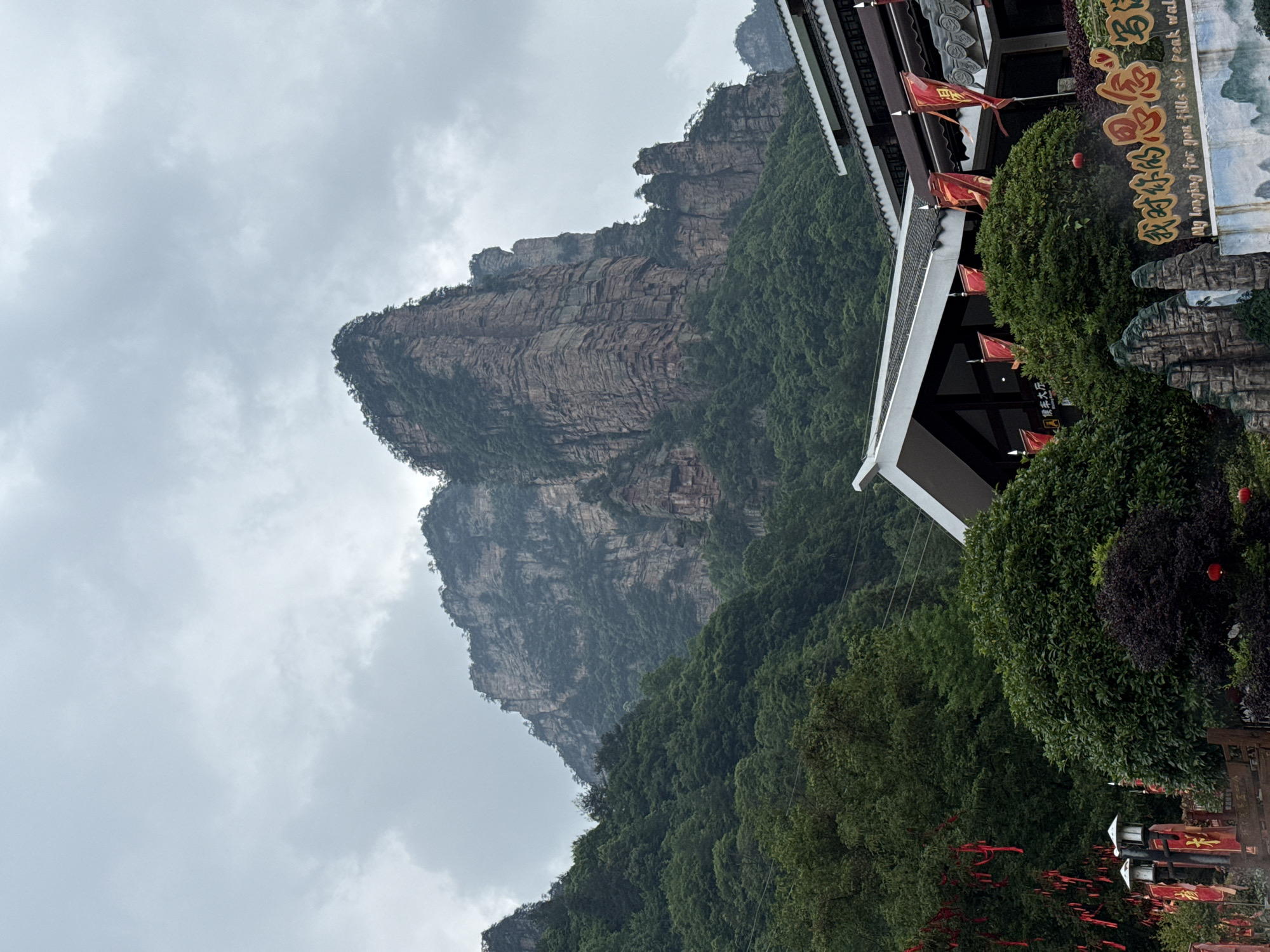The image shows a mountain in Zhonghu, China under a cloudy sky. Lush green trees cover the lower portion of the mountain. A structure with a white roof and red flags stands to the right of the mountain, partially obscured by greenery. A sign in Chinese and English reads "候车大厅 - 我对你的思念写 - My longing for you fills the peak wall." The image is oriented sideways.