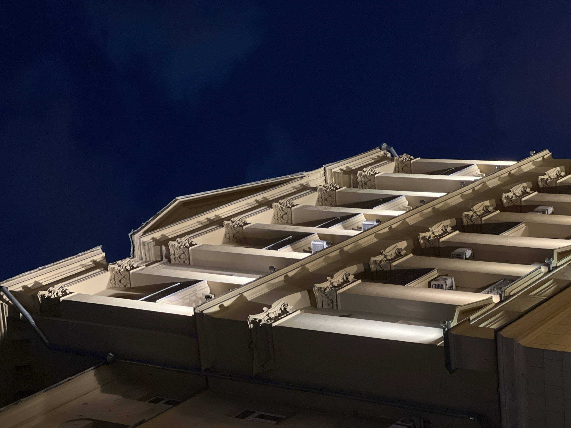 Here is a description of the image:
The image shows the exterior of a building at night. The structure has a distinct architectural style with prominent balconies and ornate details. The sky is dark blue and the building is illuminated by artificial lighting. There are no people visible.