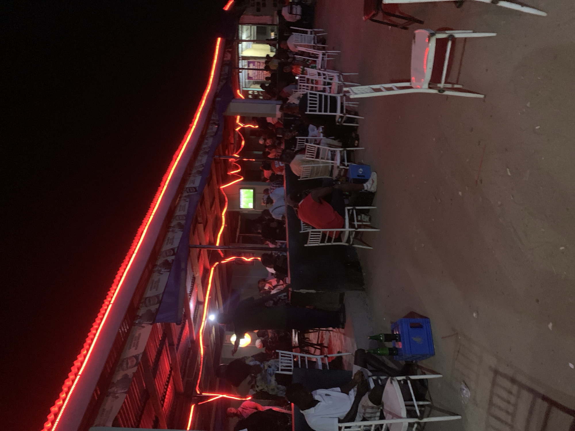 The image depicts an outdoor restaurant in Kabong, Nigeria at night. The building has a roof lined with bright red lights. People are seated at white plastic chairs and tables, some watching a television. There are bottles of beer and a blue crate visible. The ground is dirt or concrete.