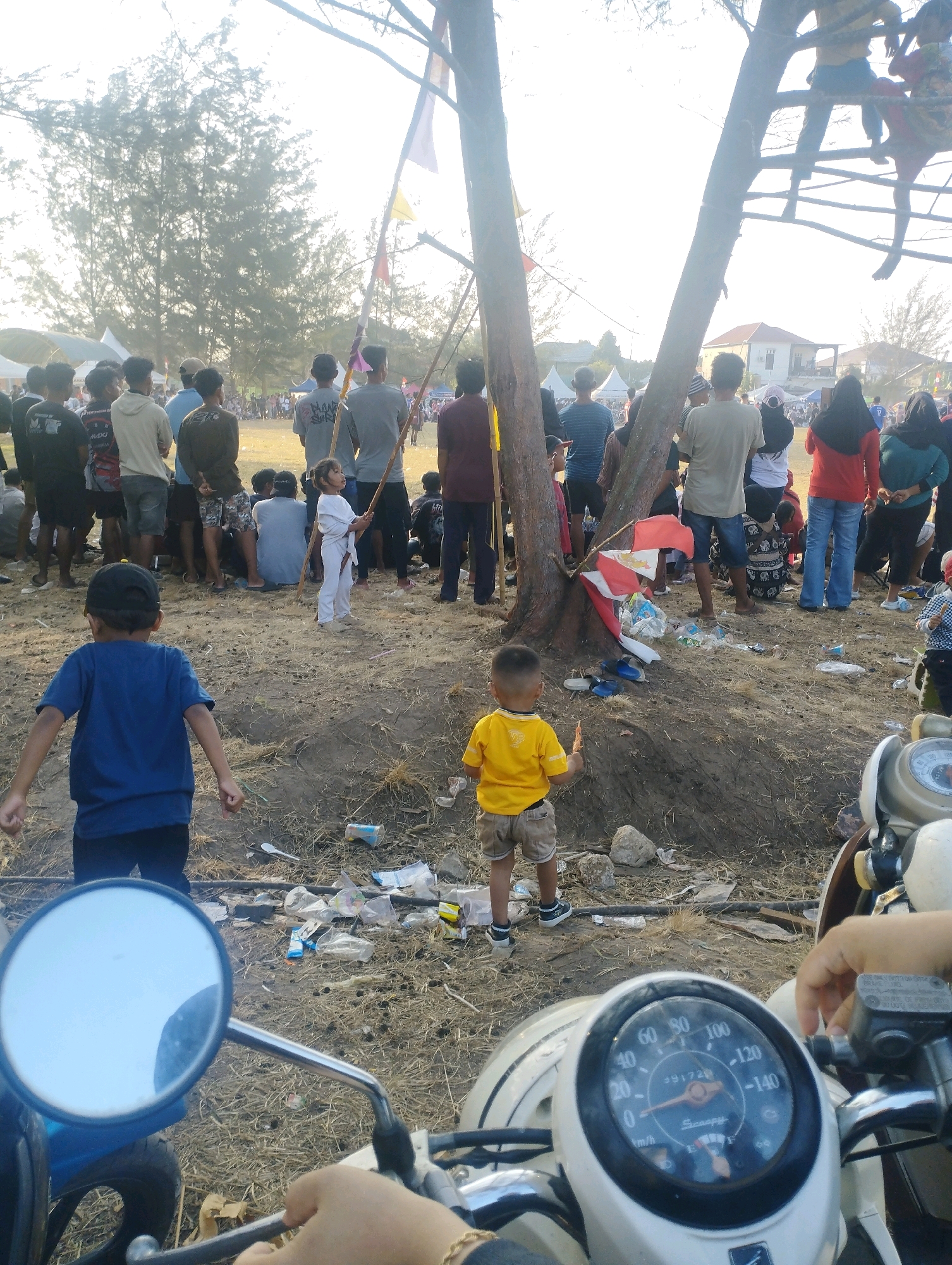 In an outdoor event setting with dry, sandy terrain, a crowd of people gathers, with some individuals standing and others seated. The atmosphere suggests a communal gathering or festival. Several children are visible, with one young boy in a blue shirt and black cap positioned in the foreground, facing away from the camera. Another child, wearing a yellow shirt and shorts, stands further back, also facing away. A young girl in a white uniform, possibly a martial arts gi, stands near a tree trunk holding a stick.
The environment is characterized by sparse vegetation and scattered litter, including plastic bottles and wrappers. A prominent tree with multiple branches is a central element, with some people, including a child, perched on its limbs. Flags of various colors are displayed, suggesting a festive or celebratory occasion. In the foreground, the handlebars and dashboard of a scooter are visible, with a hand holding onto the handlebars, indicating the perspective of someone on or near the vehicle. The sky is bright, suggesting daylight, possibly late afternoon or early morning given the lighting. The overall scene depicts a lively outdoor event with a mix of people of different ages.