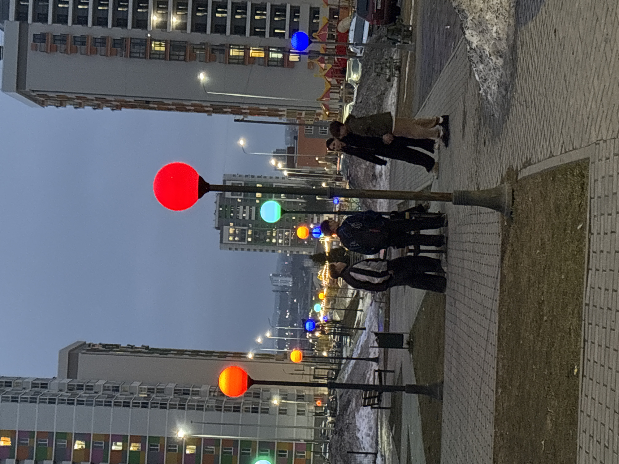 The image shows a street in Izhevsk, Russia, at dusk. The street is lined with tall apartment buildings on both sides and features a row of decorative streetlights with brightly colored spherical lamps (red, orange, blue, and green). Several people are walking along the sidewalk. The ground is paved with grey interlocking bricks. Patches of snow are visible along the edge of the sidewalk. A playground can be seen in the background, beyond the closest building.